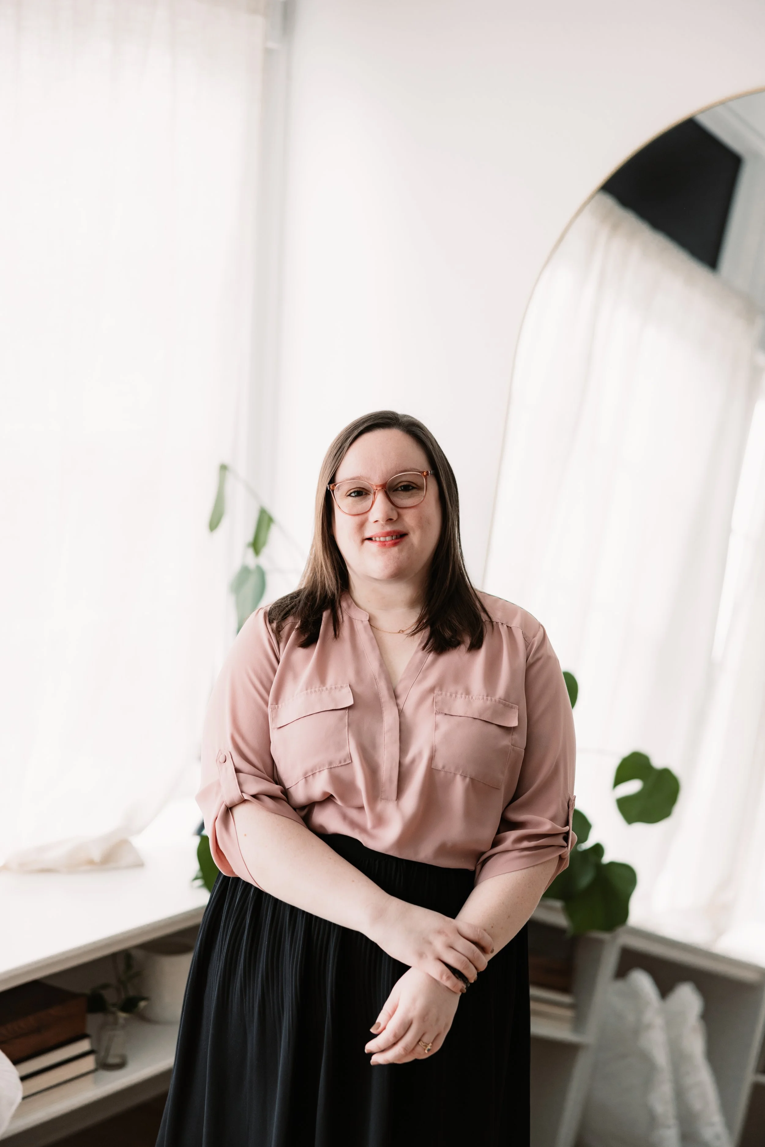 A woman with long brown hair, wearing glasses and a black top, smiling and taking a selfie in a room with beige walls.