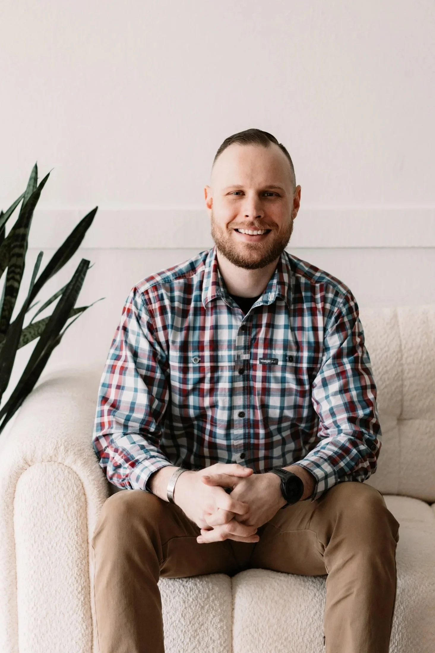Portrait of a smiling man with short dark hair, wearing a navy blue Under Armour jacket over a plaid shirt, standing against a plain off-white wall.