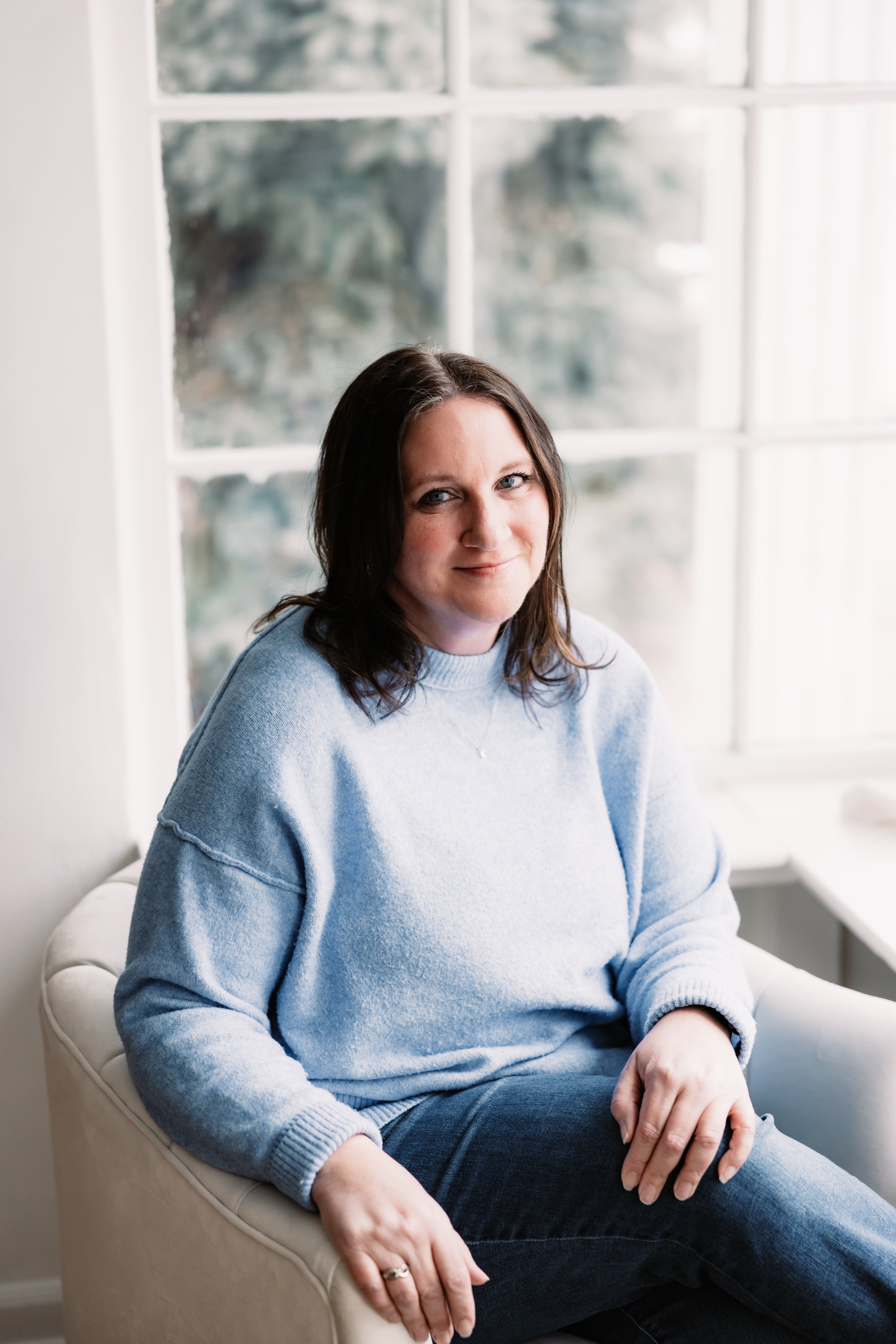 A woman with wavy brown hair and fair skin smiling at the camera, wearing a beige coat, sitting against a plain light-colored wall.