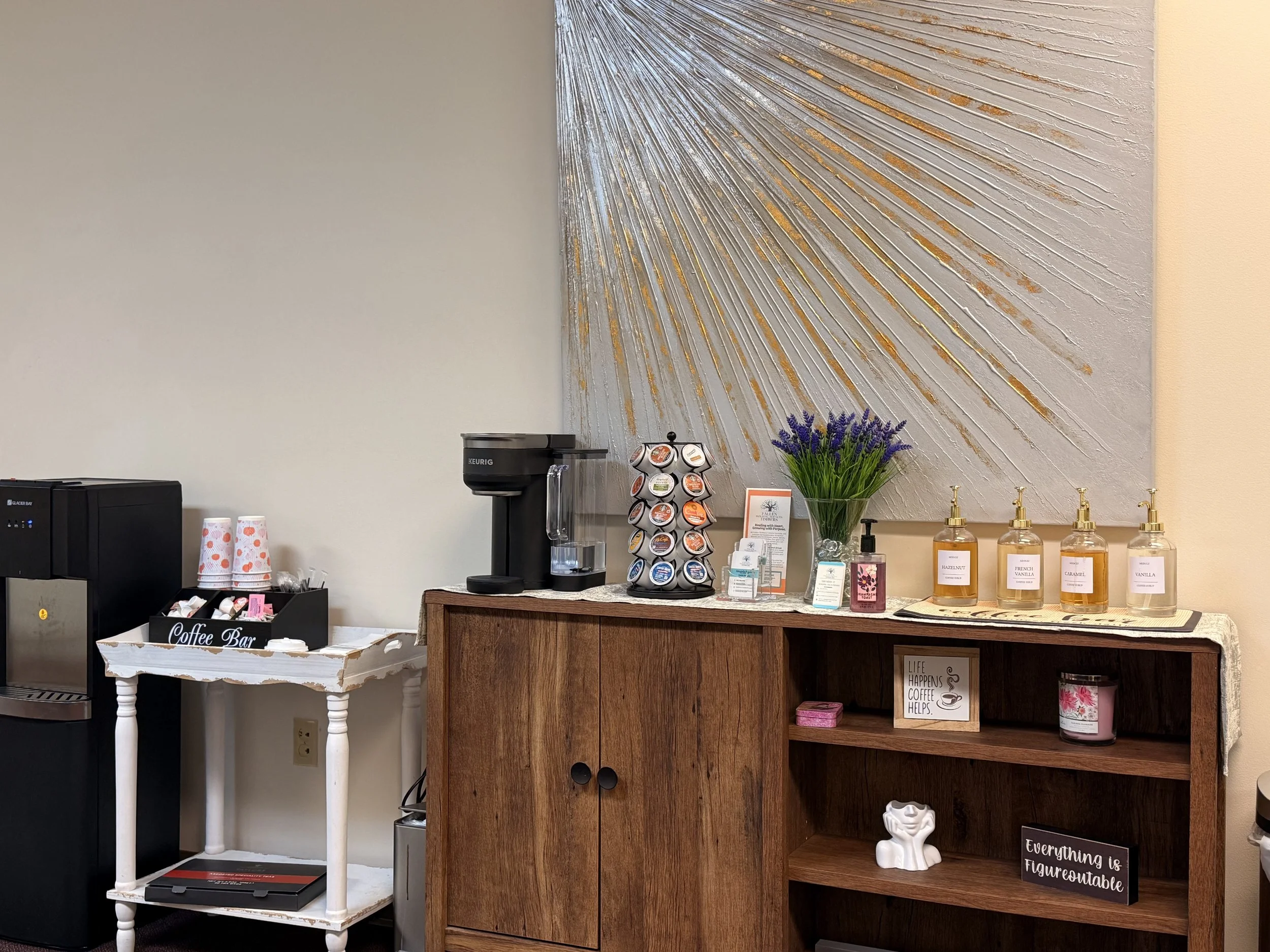 Coffee station with a black coffee maker, a coffee pods display, and various flavored syrups on a wooden sideboard, decorated with lavender in a clear vase and wall art with gold accents.