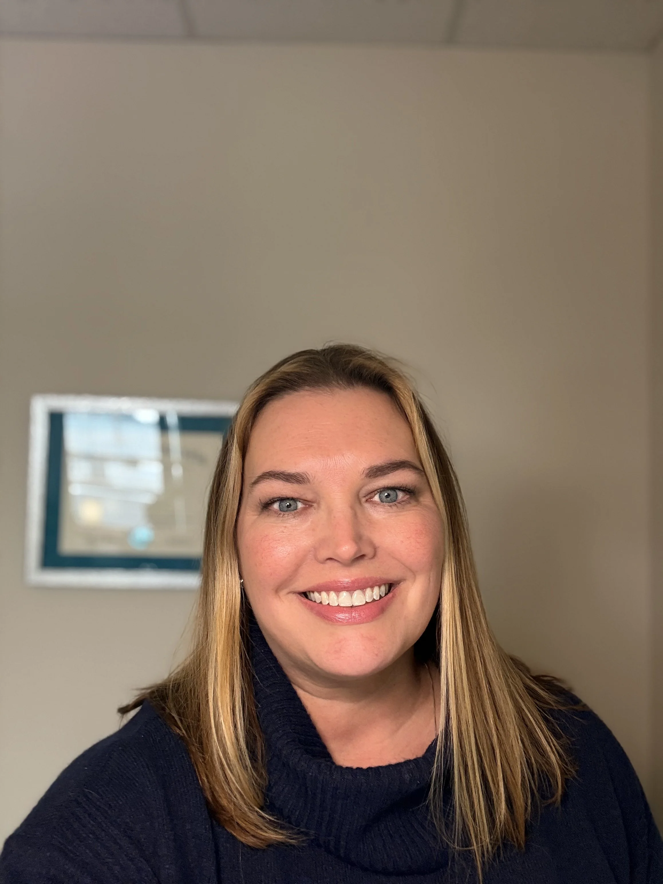 A woman with blonde hair and a blue sweater smiling indoors with a framed picture on the wall in the background.