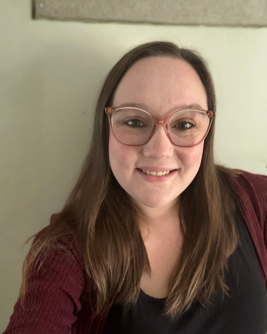 A woman with long brown hair, wearing pink glasses, smiling at the camera, with a light-colored background and a maroon sweater.