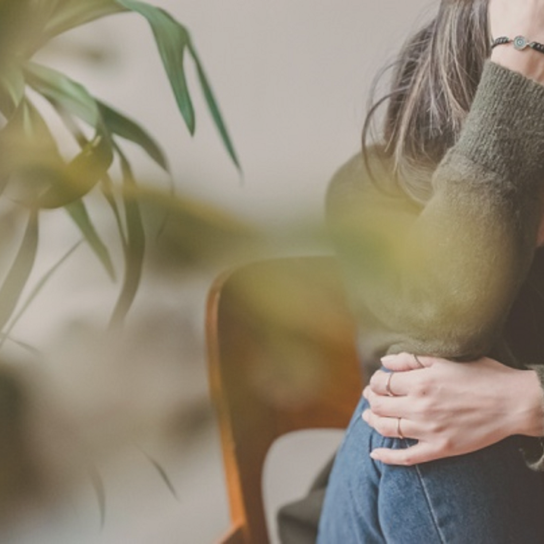 A woman sitting at a desk with a large green plant in the foreground. The woman is wearing an olive green sweater and blue jeans, with her arm resting on her knee and her hand on her stomach.