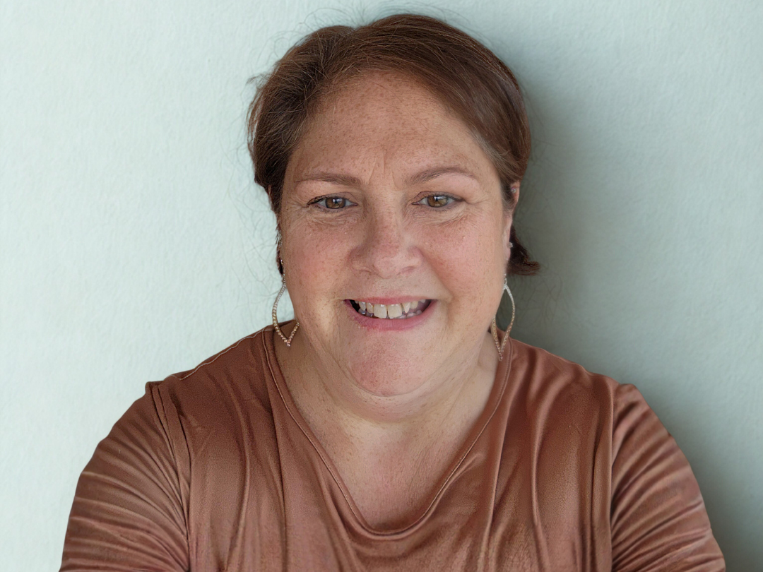 Close-up of a woman with reddish-brown hair, smiling, wearing hoop earrings and a brown satin top, standing against a light green wall.