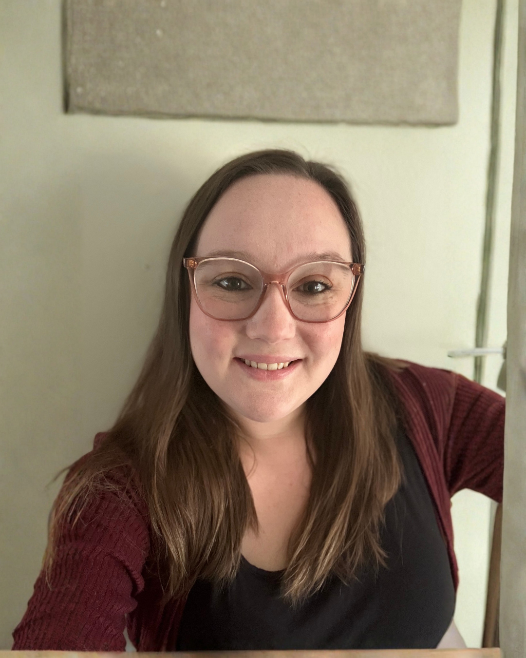 A woman with long brown hair, wearing glasses and a black top, smiling and taking a selfie in a room with beige walls.