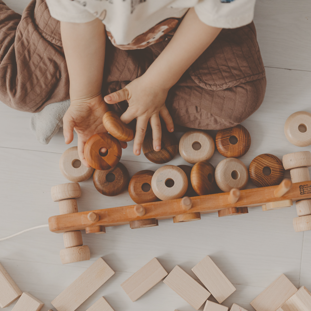 Child playing with wooden stacking toys and blocks on a light-colored wooden floor.