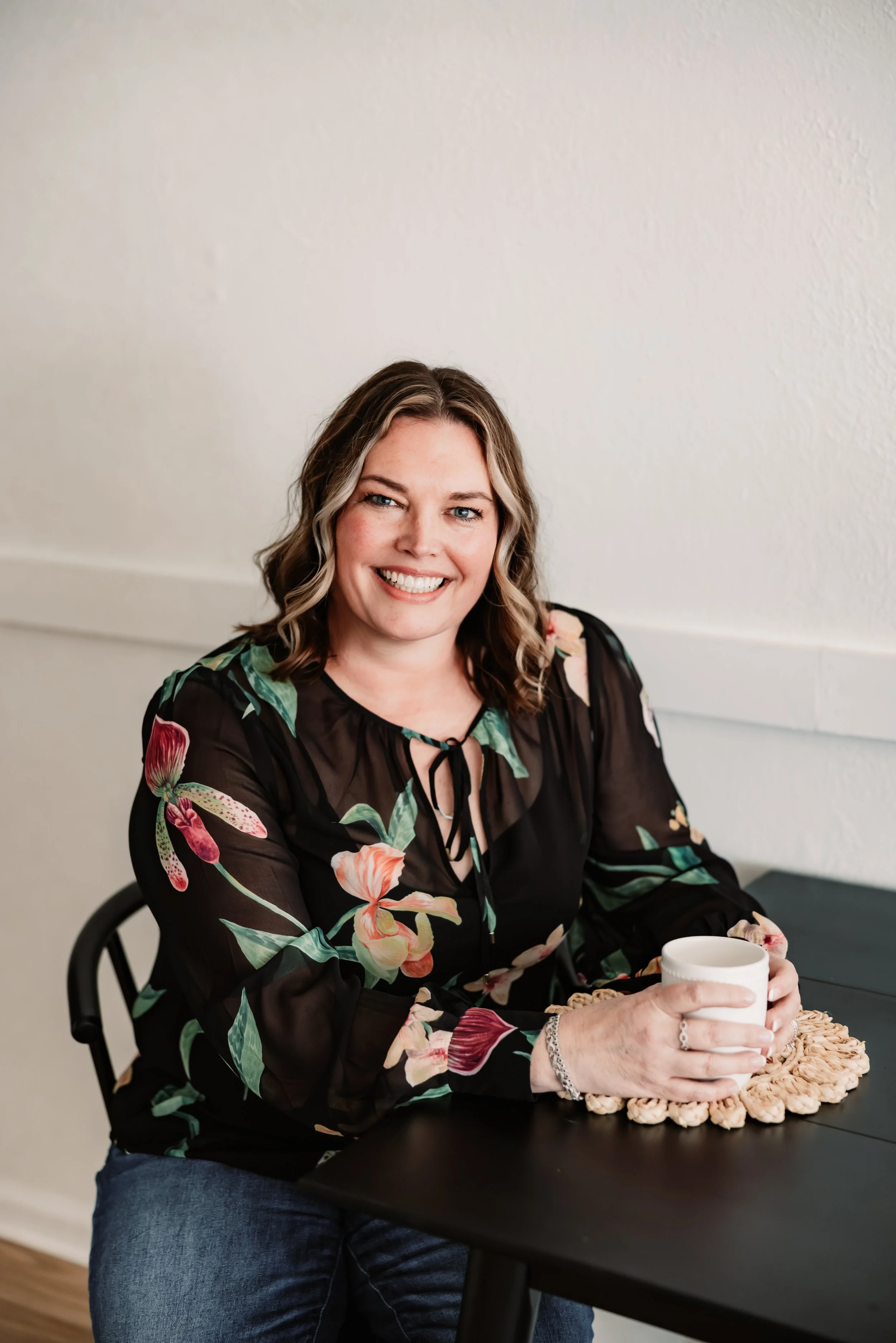 A woman with reddish-brown hair and a green sweater sitting in a black office chair, smiling at the camera, in a room with gray walls, a white door, and a bookshelf with colorful books and a potted plant.