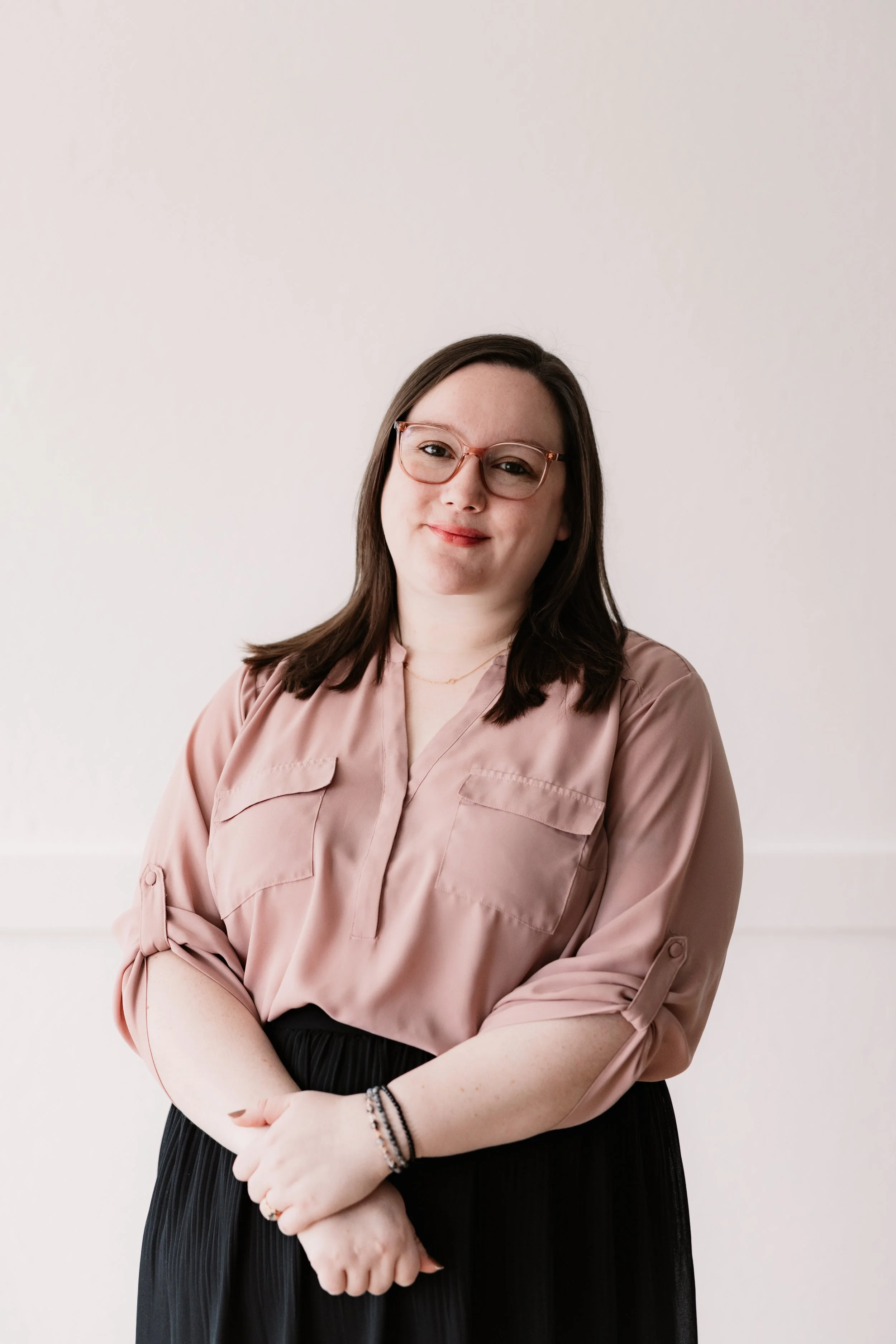 A woman with long brown hair, wearing pink glasses, smiling at the camera, with a light-colored background and a maroon sweater.