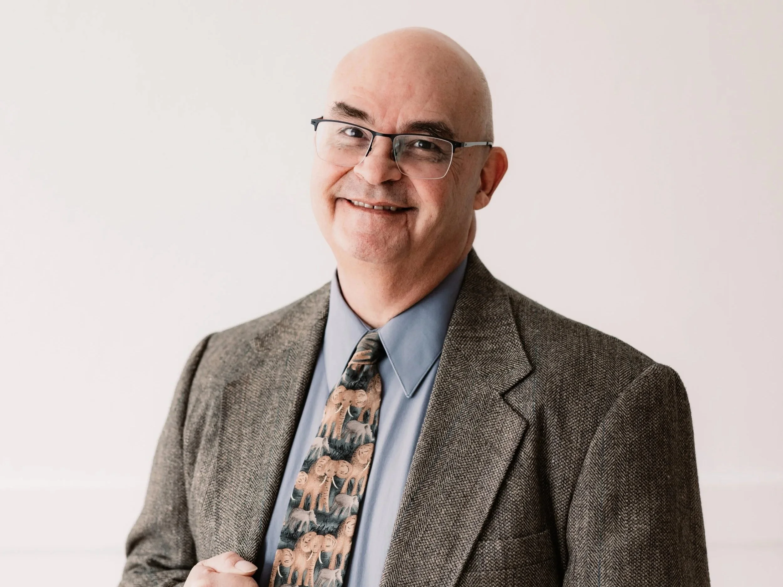 smiling bald man with glasses wearing a cream-colored knit sweater, indoor background with blurred wall decorations