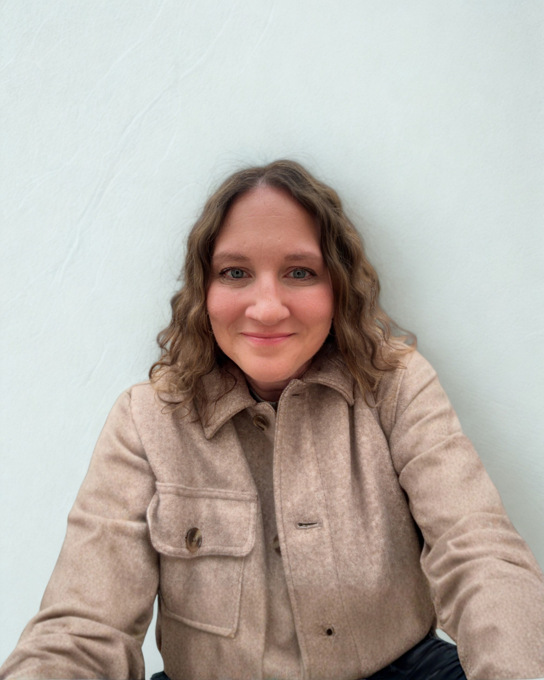 A woman with wavy brown hair and fair skin smiling at the camera, wearing a beige coat, sitting against a plain light-colored wall.