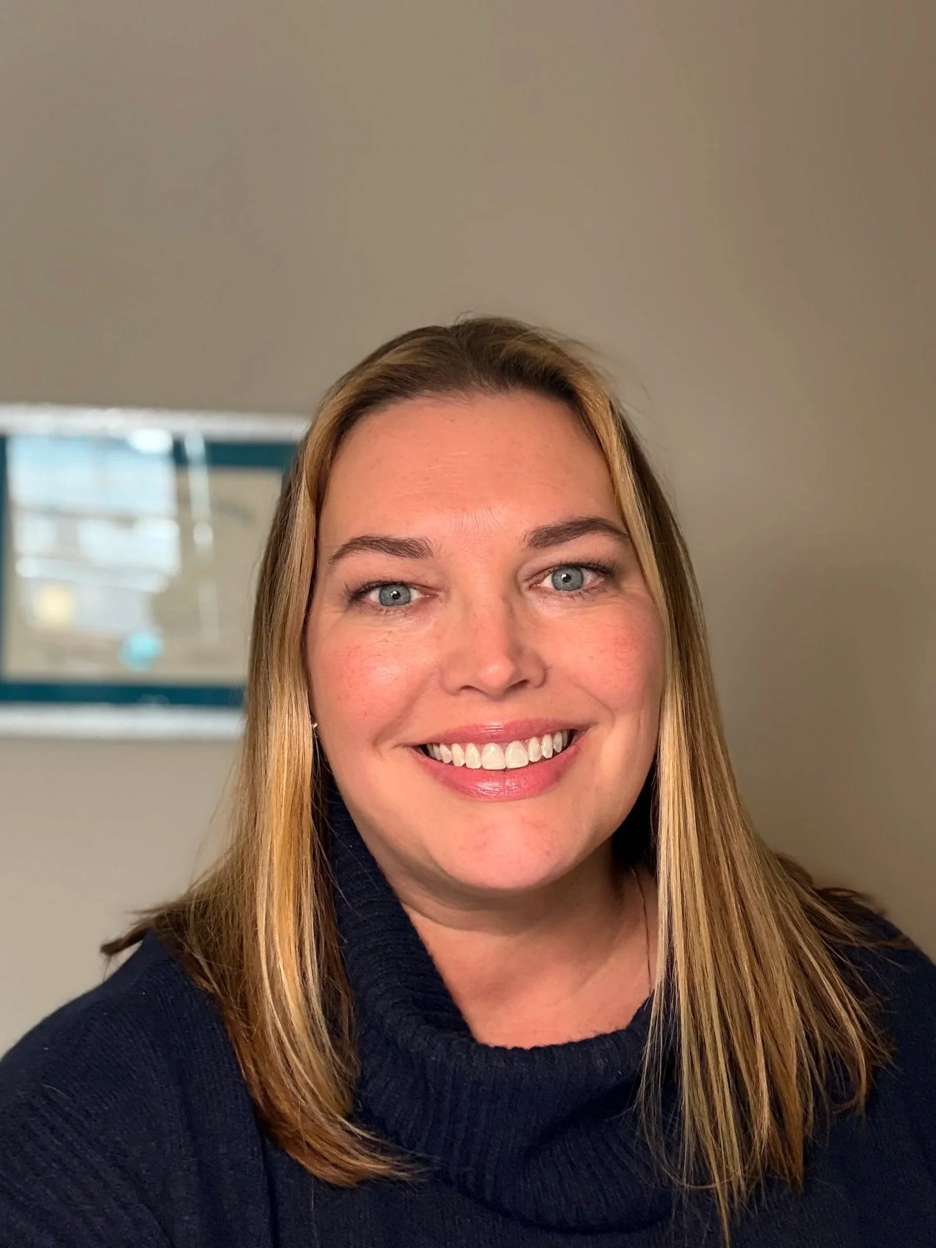A woman with reddish-brown hair and a green sweater sitting in a black office chair, smiling at the camera, in a room with gray walls, a white door, and a bookshelf with colorful books and a potted plant.