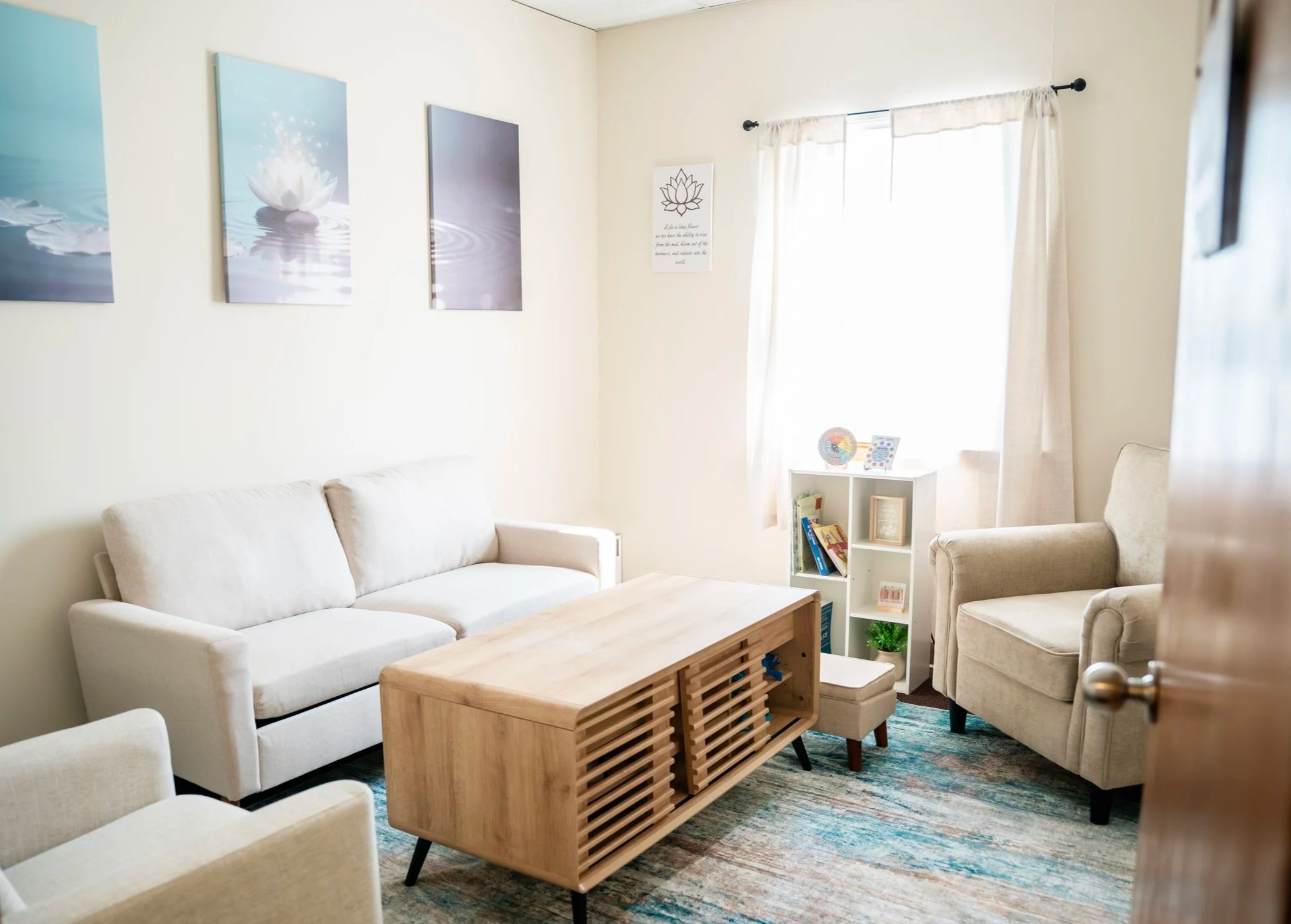 A cozy living room with white sofa chairs, a wooden coffee table, a small white shelving unit with books and decorative items, and wall art including a Lotus flower. There is a window with sheer curtains allowing natural light to brighten the room, and a colorful area rug on the floor.