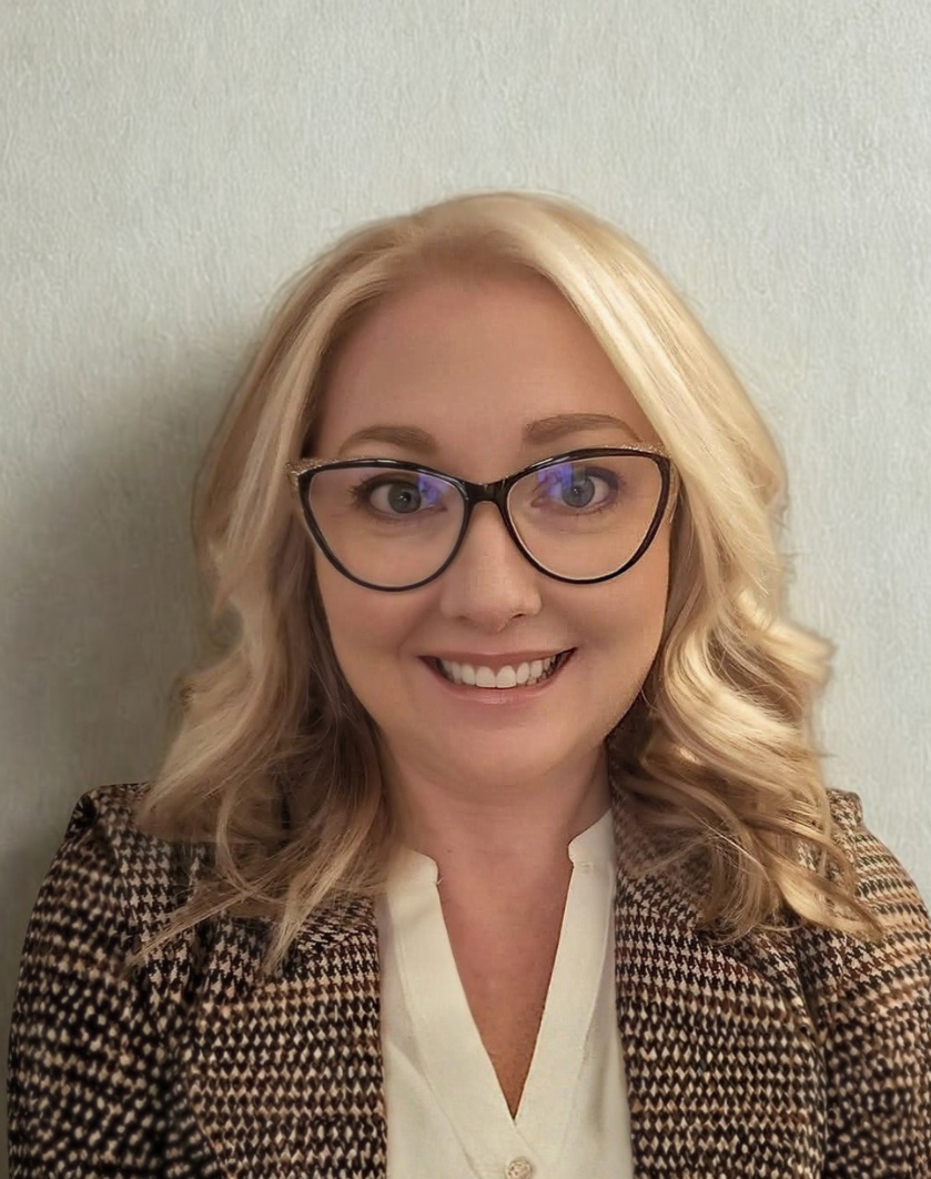 A woman with blonde, wavy hair wearing glasses, a patterned blazer, and a white blouse, smiling at the camera against a plain light-colored wall.