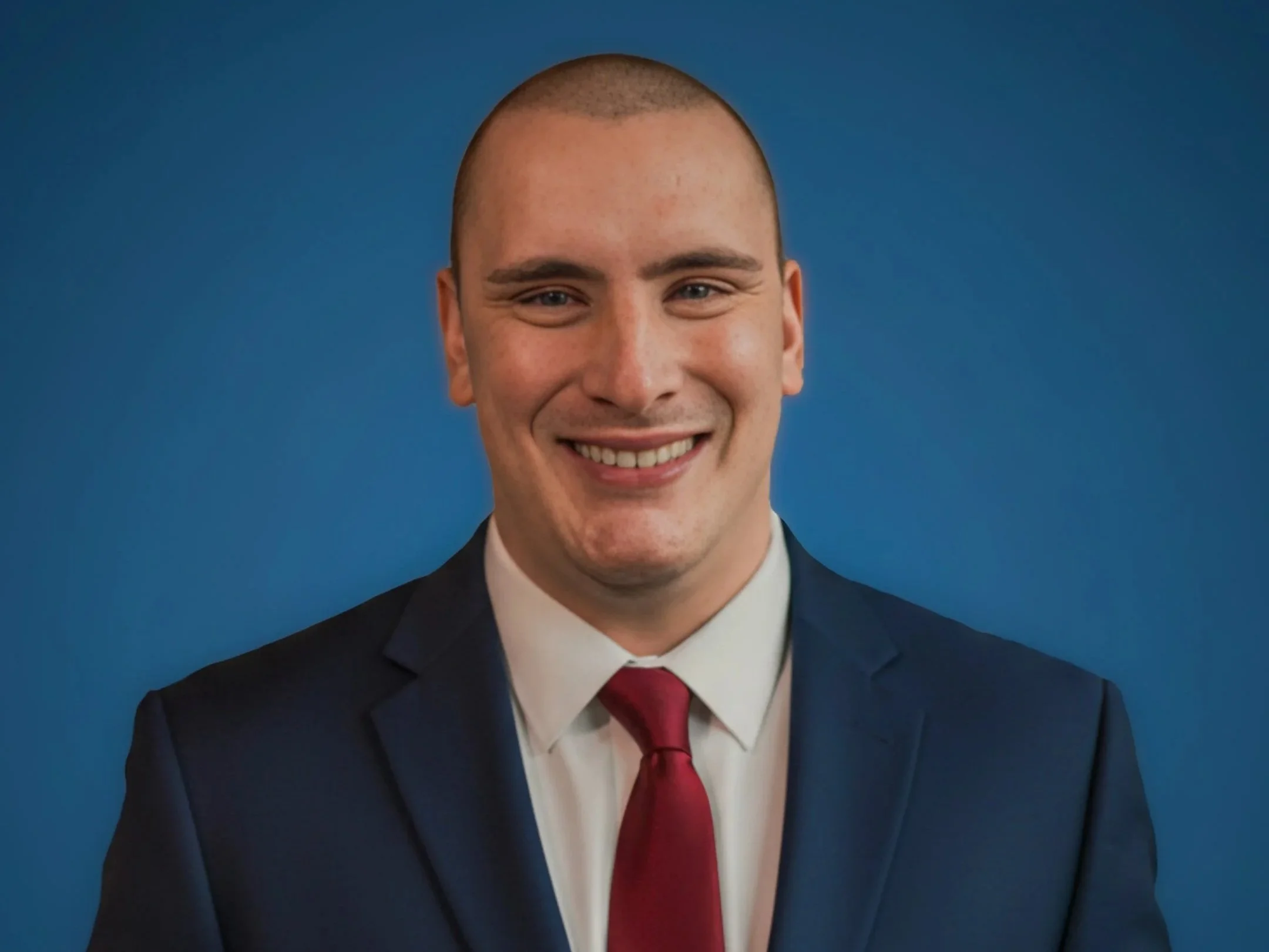 A young man in a dark suit, white shirt, and red tie, smiling against a blue background.