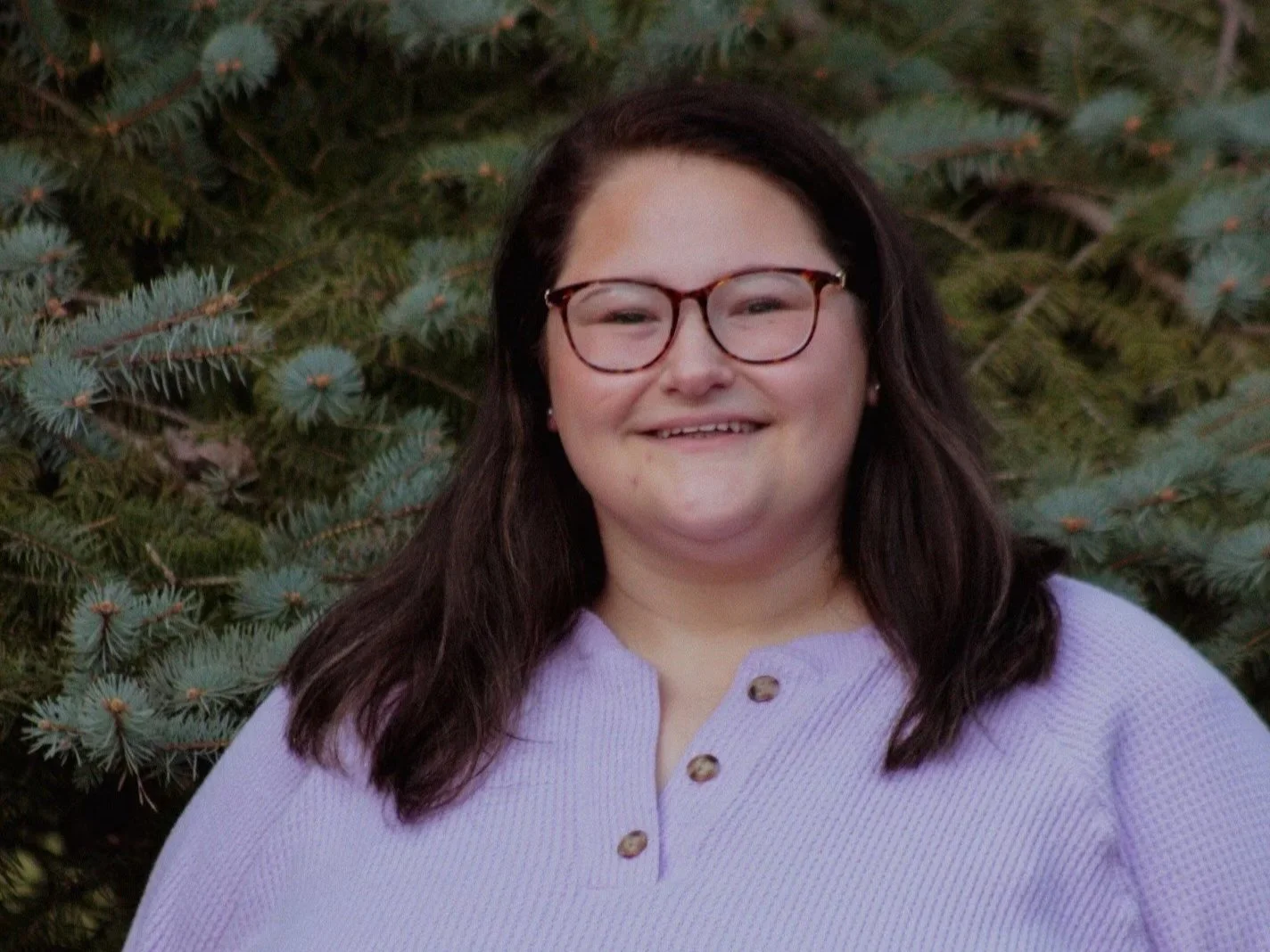 A woman with dark hair, glasses, and a light purple top, smiling in front of a pine tree.