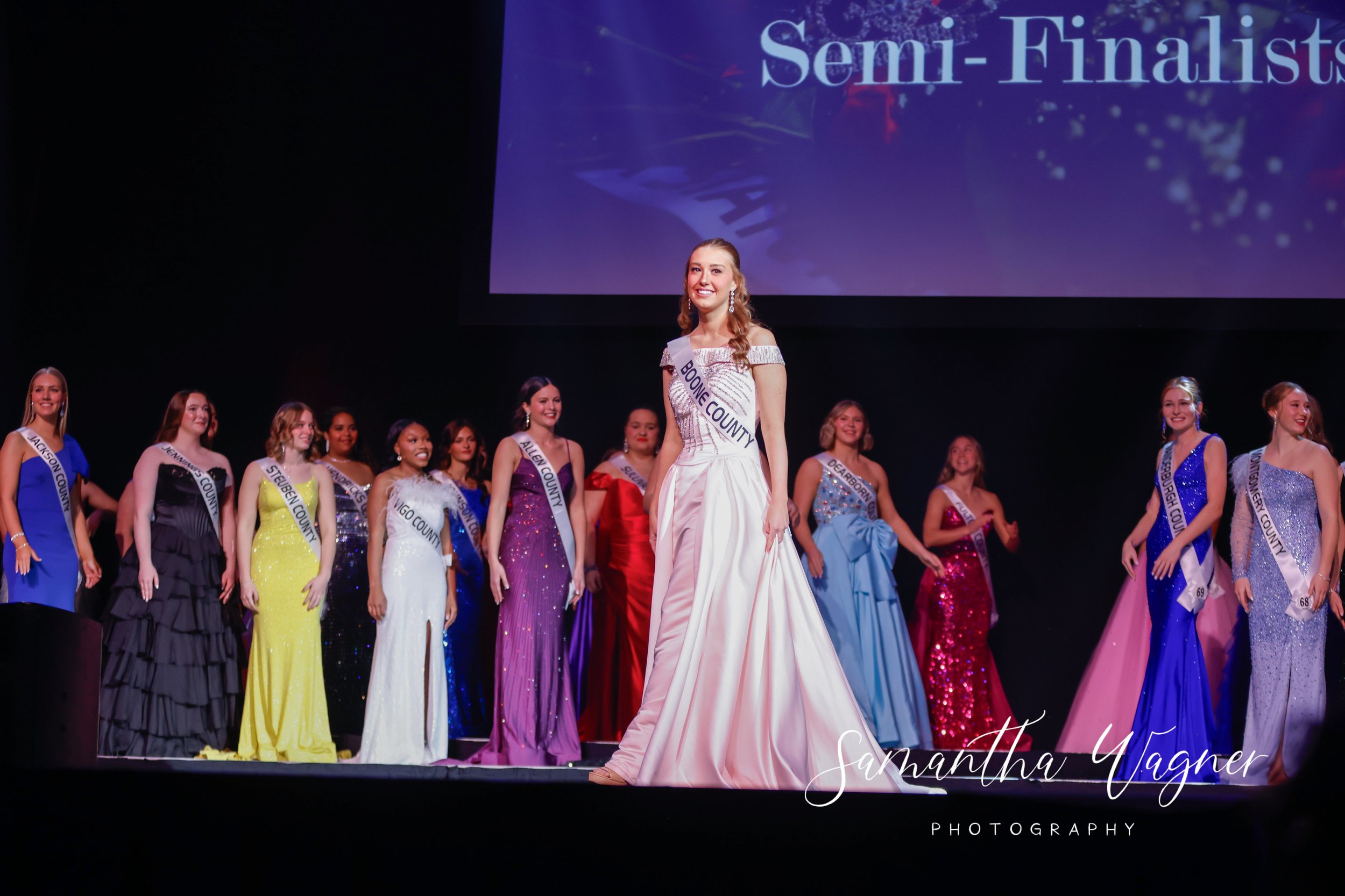Pageant contestants on stage, wearing evening gowns and sashes, with the text 'Semi-Finalists' displayed above.
