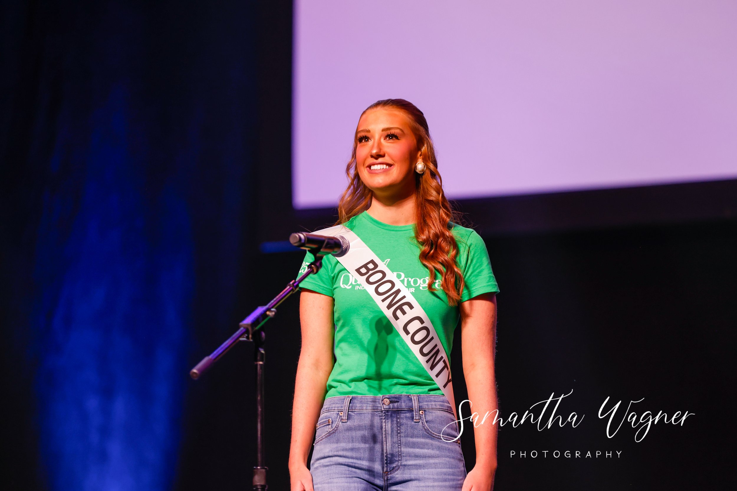 Person wearing a Boone County sash and green shirt, standing at a microphone on stage, with a dark background.