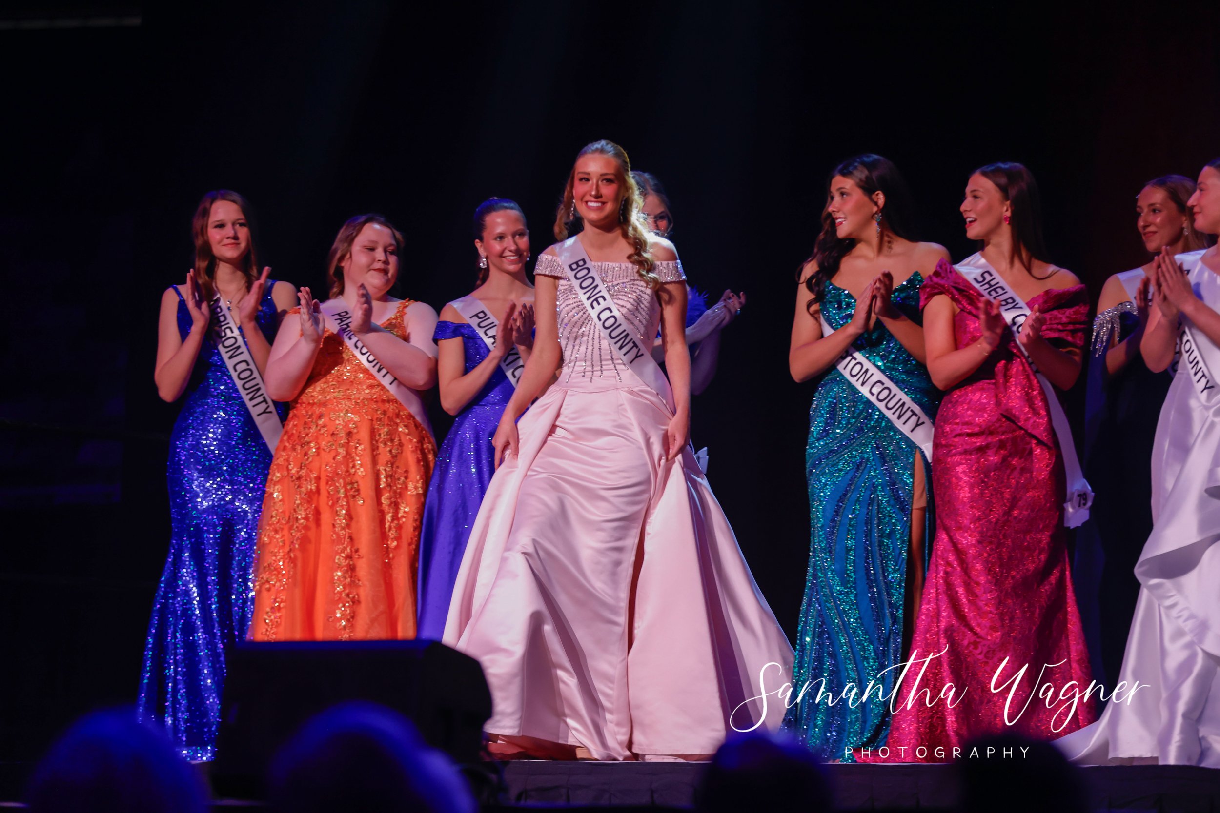 Beauty pageant contestants in gowns, sashes displaying county names, on stage during award ceremony.