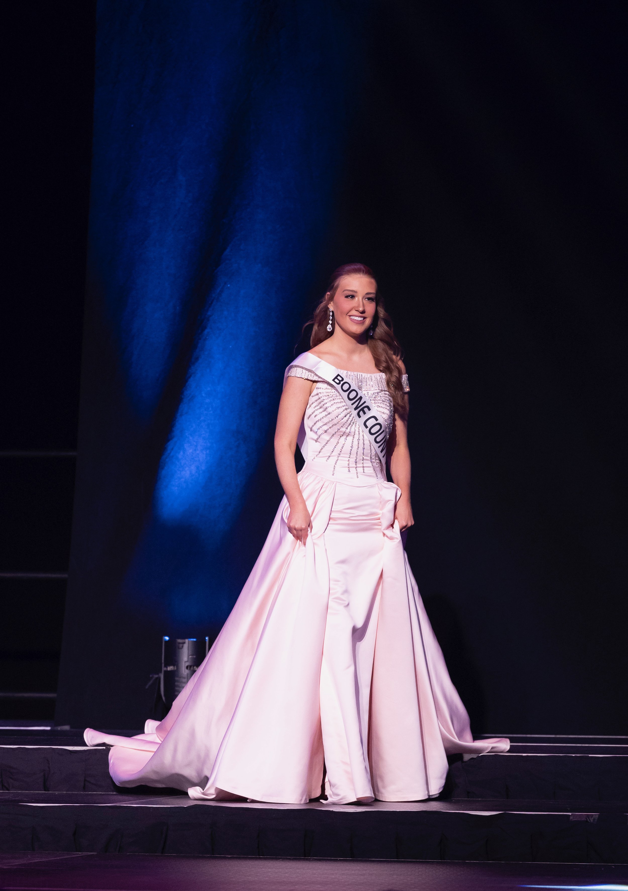 Pageant contestant wearing a white gown with "Boone County" sash on stage.