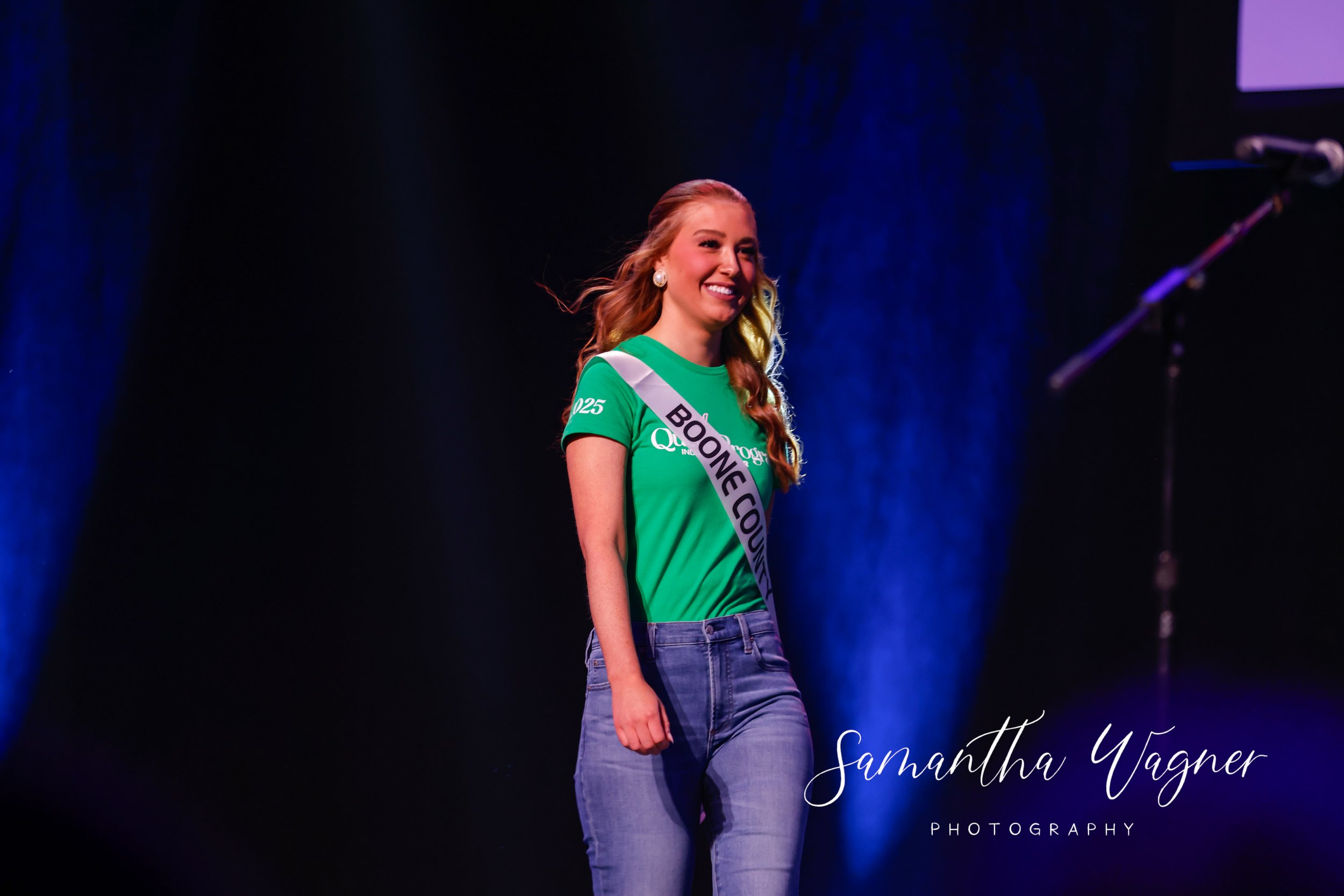 Person on stage with "Boone County" sash, wearing a green t-shirt and jeans, smiling under stage lights.