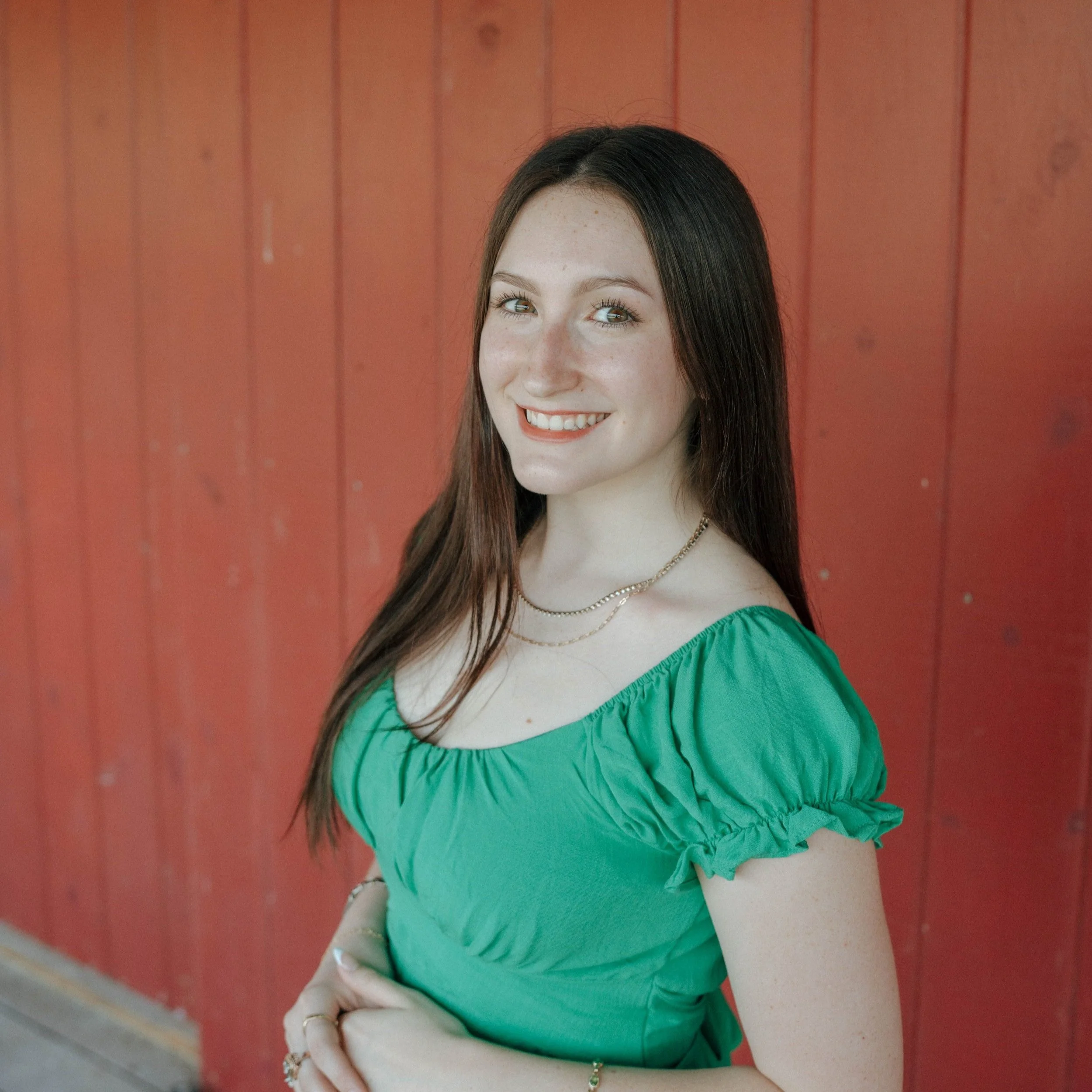 Portrait of a woman with blonde hair, wearing a pearl necklace and light-colored top, smiling. Background includes a kitchen area with wooden cabinets.