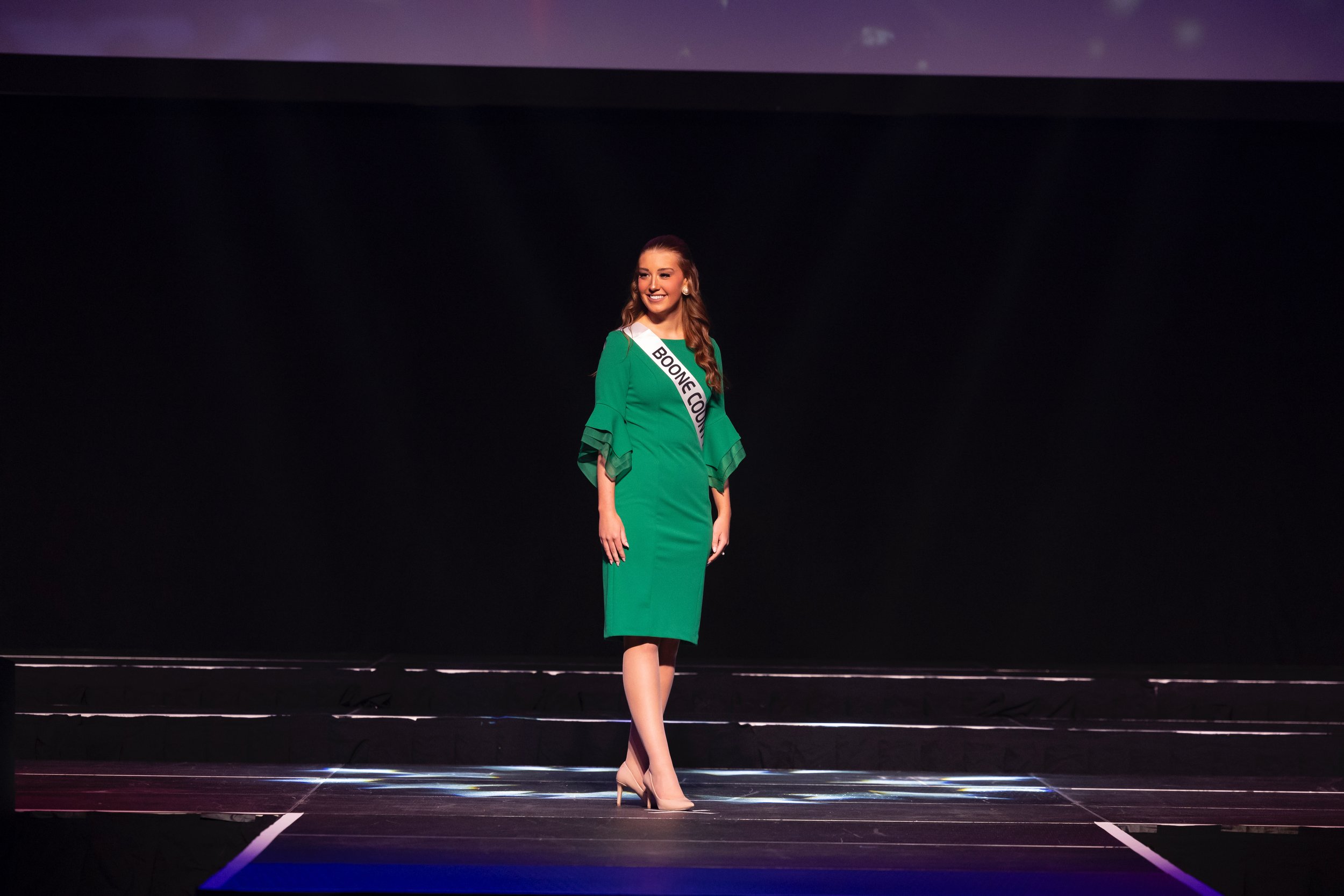 Woman in green dress wearing a sash labeled 'Boone County' on a stage.