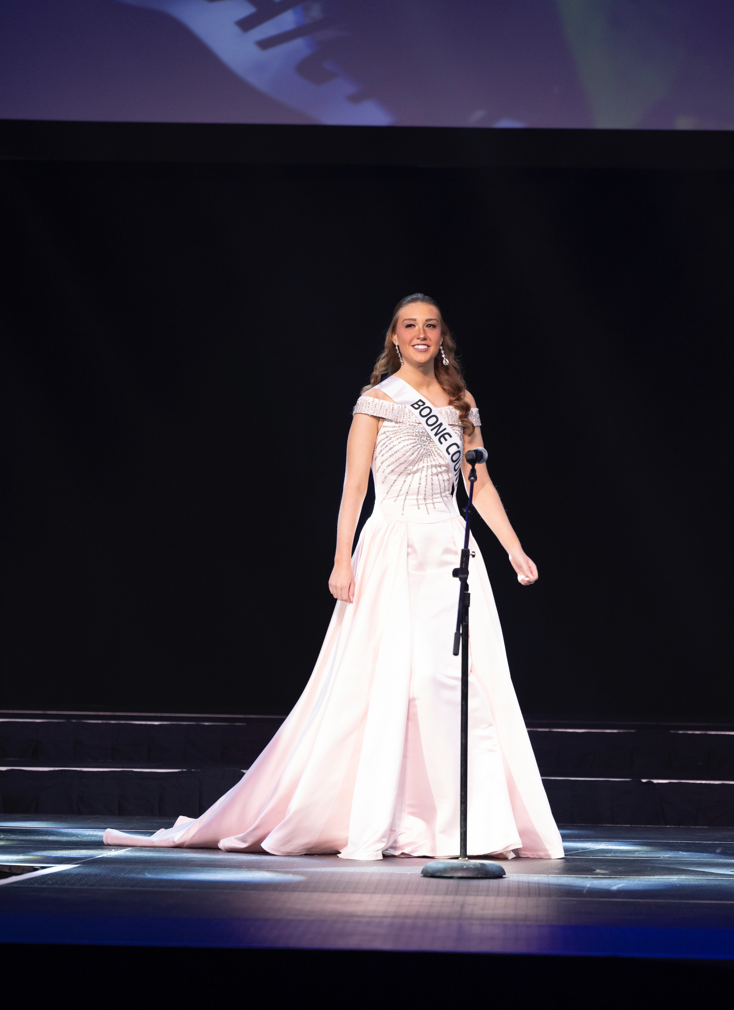 Pageant contestant on stage in a formal gown with a sash labeled 'Boone Co' standing before a microphone.