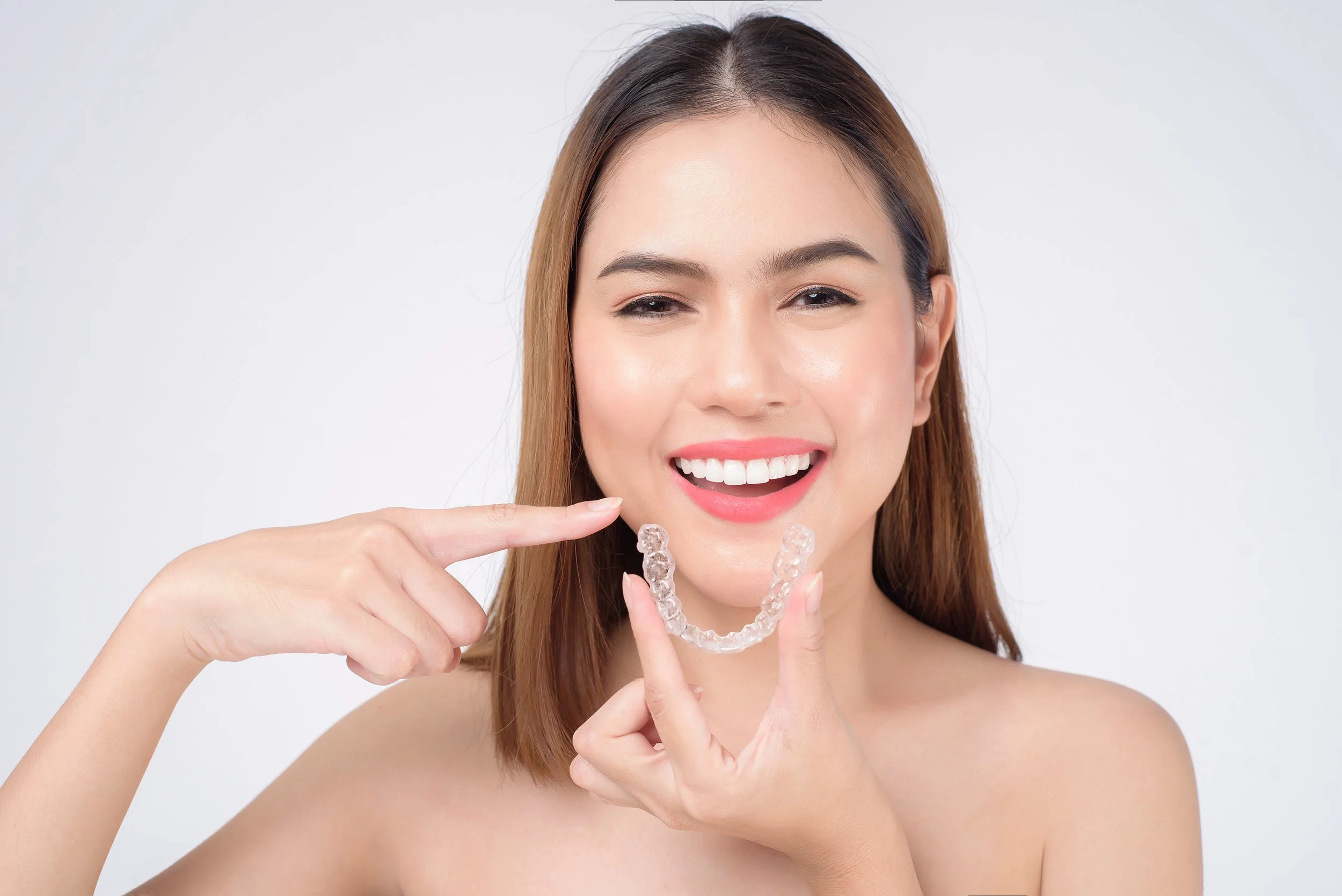 A woman holding and pointing to a clear dental aligner, smiling and winking.