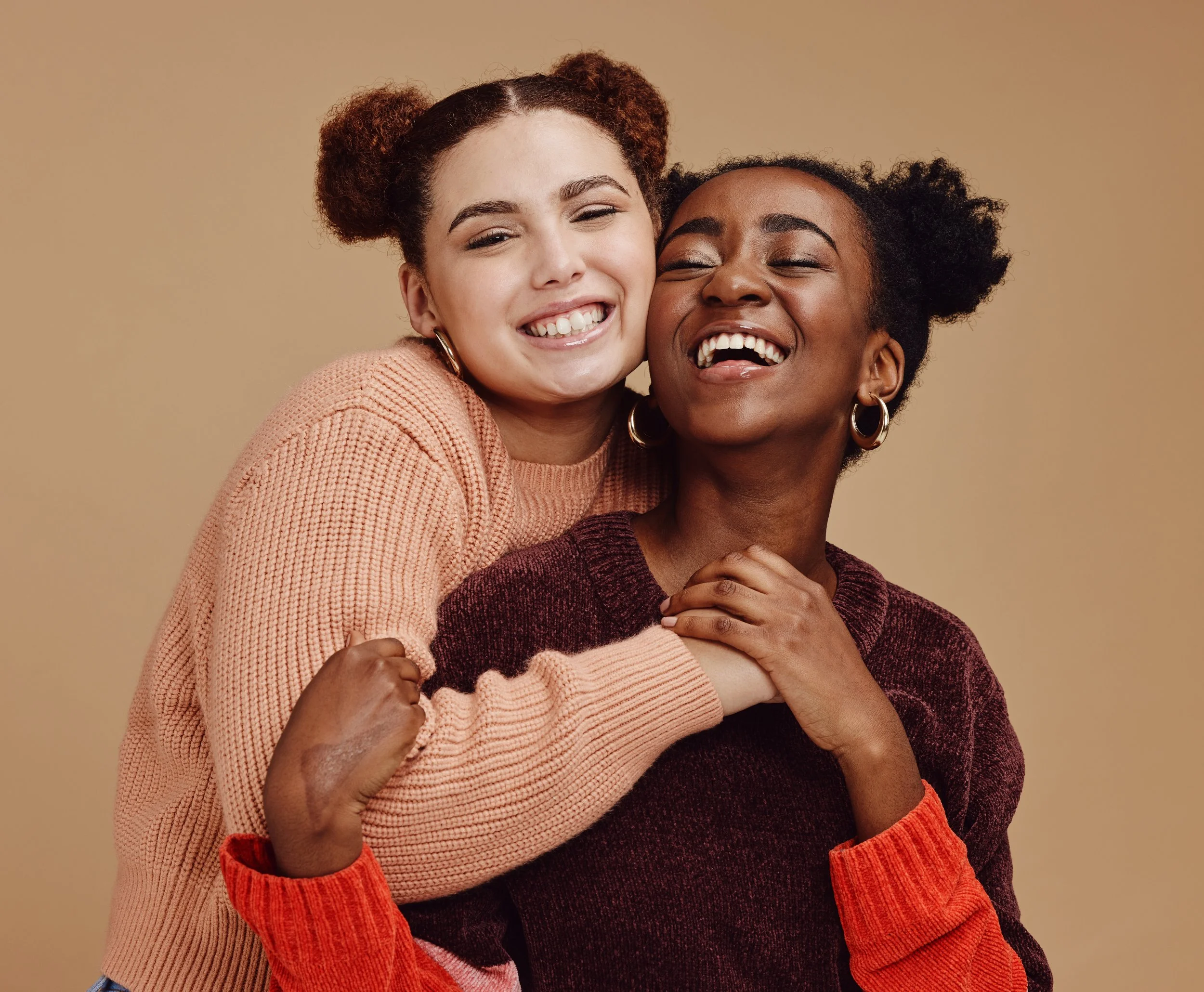Two women smiling and hugging each other against a beige background. One woman has light skin, brown curly hair styled in puffs, and is wearing a peach-colored sweater. The other woman has dark skin, black curly hair styled in puffs, and is wearing a dark maroon sweater with orange cuffs.