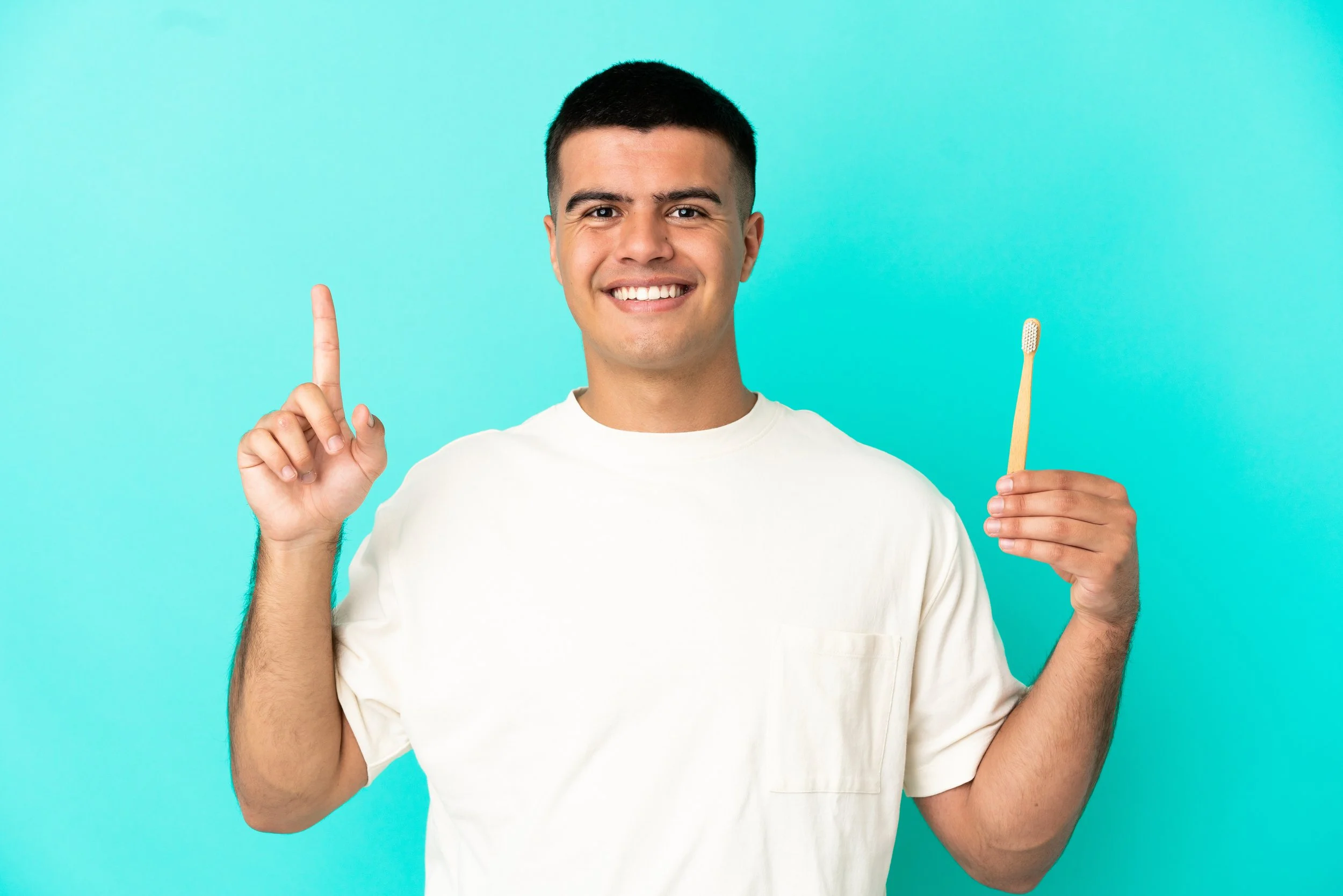 Young man smiling while holding a toothbrush in his right hand, pointing upward with his left index finger, against a bright blue background.