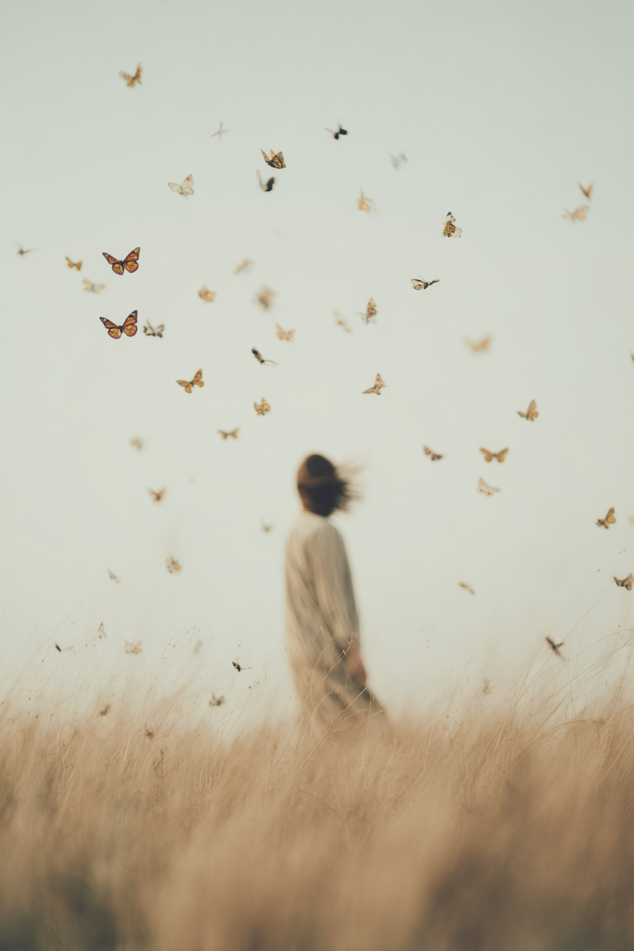 A person with shoulder-length hair wearing a light-colored dress is standing in a field of tall, dry grass with their back turned and head slightly bent. Butterflies are flying around and above them against a pale sky.