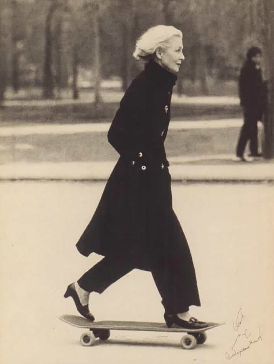 An elderly woman riding a skateboard in a park, dressed in a long coat and heels.