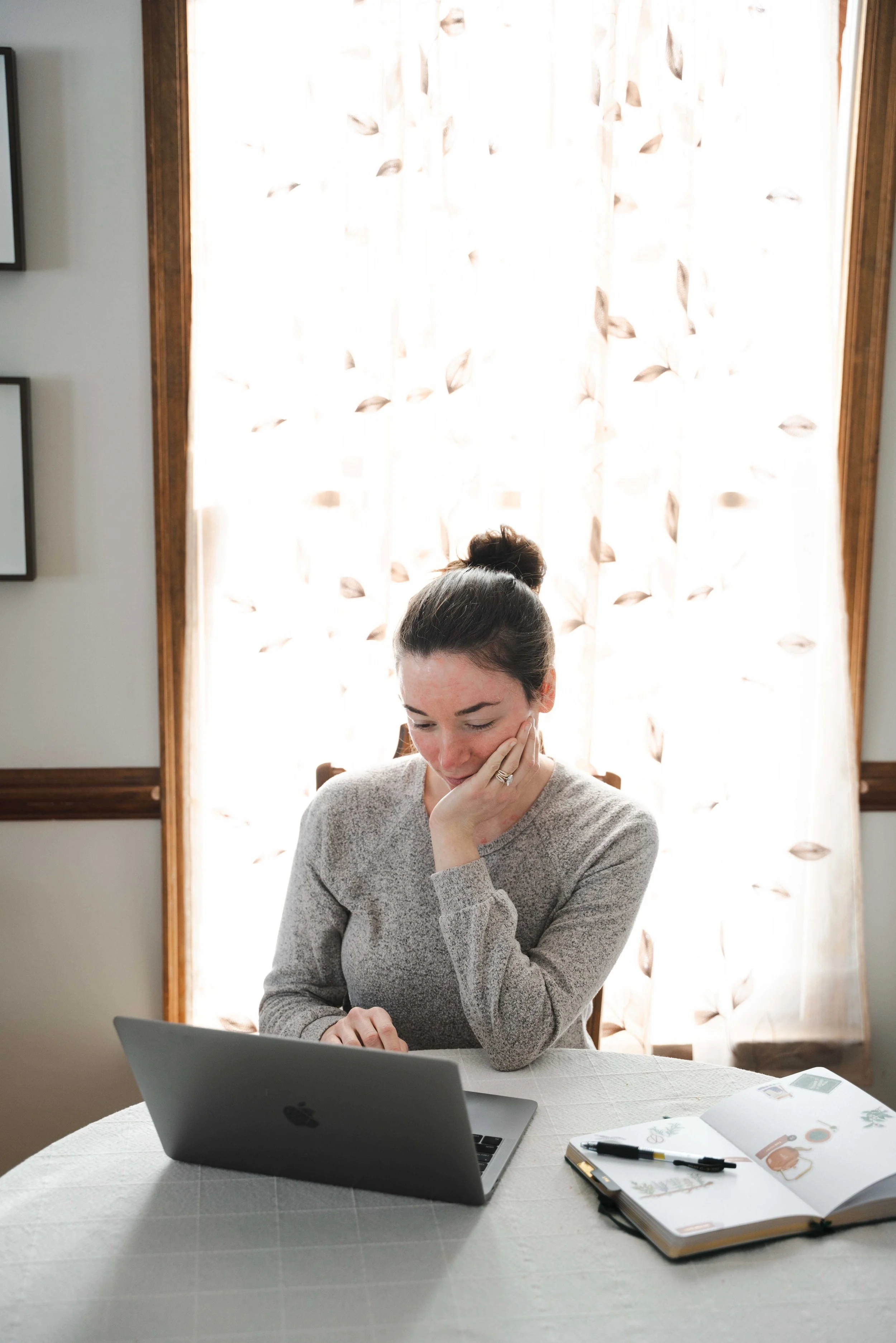 A woman sitting at a table looking at a laptop with a thoughtful expression, resting her chin on her hand, in a room with floral curtains, an open planner, and a pen on the table.