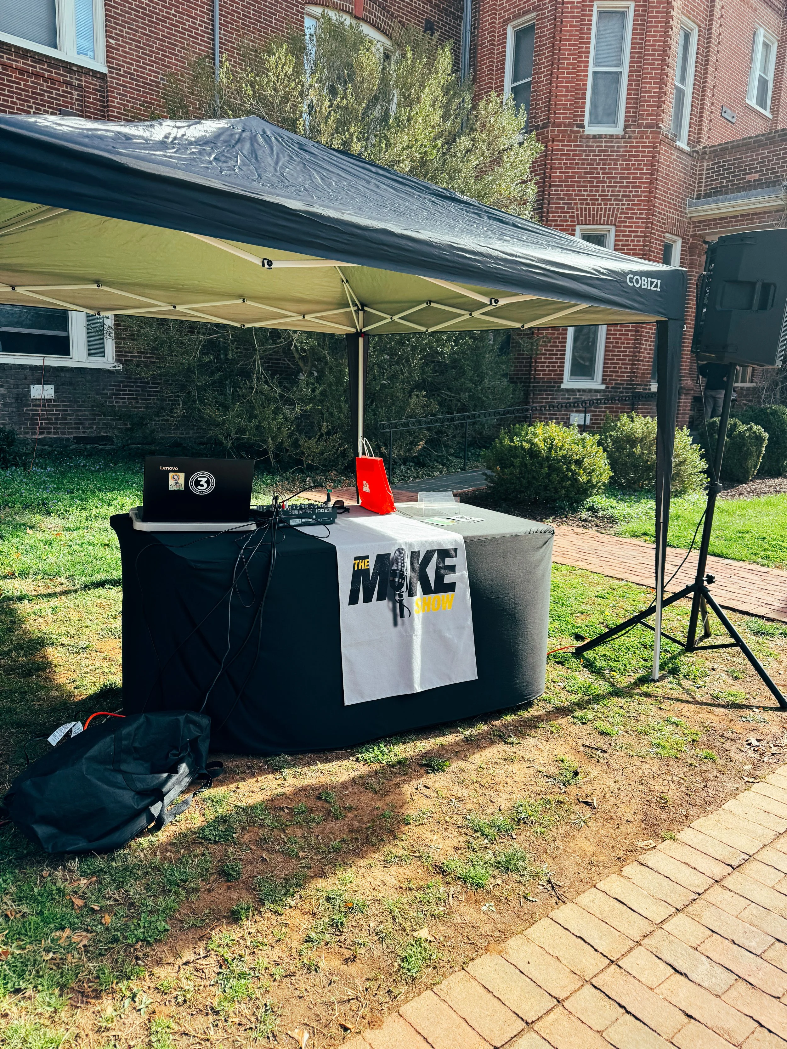 An outdoor scene with a black canopy tent and a table underneath, featuring a banner that says 'The Make Show' with a microphone logo. There is a laptop and audio equipment on the table. Surrounding the scene are brick buildings, greenery, and a paved walkway.