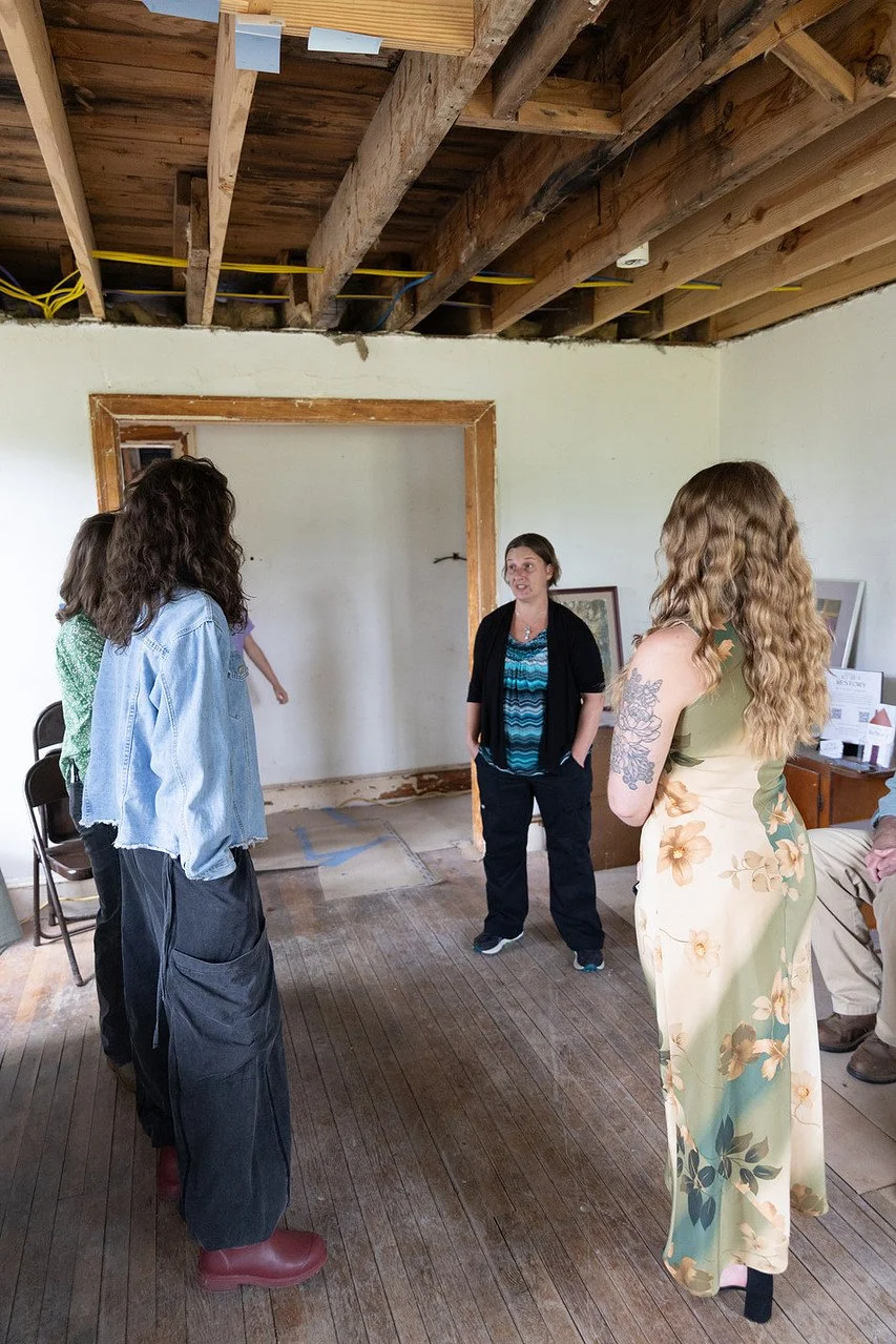 Group of women in a partially renovated room with exposed wooden ceiling beams, standing and talking, with framed pictures and construction materials in the background.