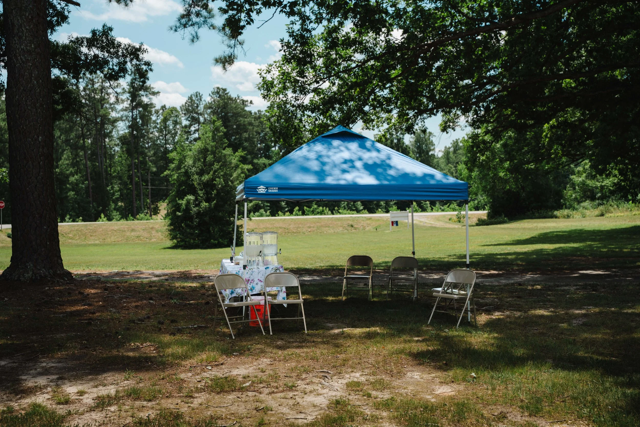 An outdoor setup with a blue canopy tent, chairs, and a table with drinks, surrounded by trees on a grassy area under a partly cloudy sky.