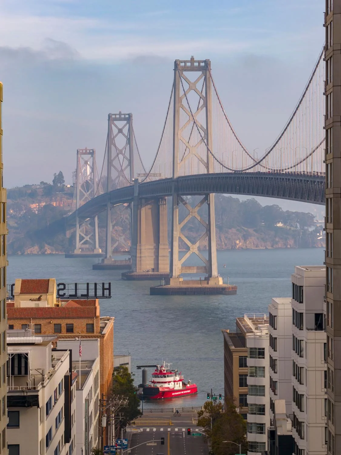 View of the San Francisco Bay Bridge seen through city buildings, with a red boat and water in the foreground and hills in the background.