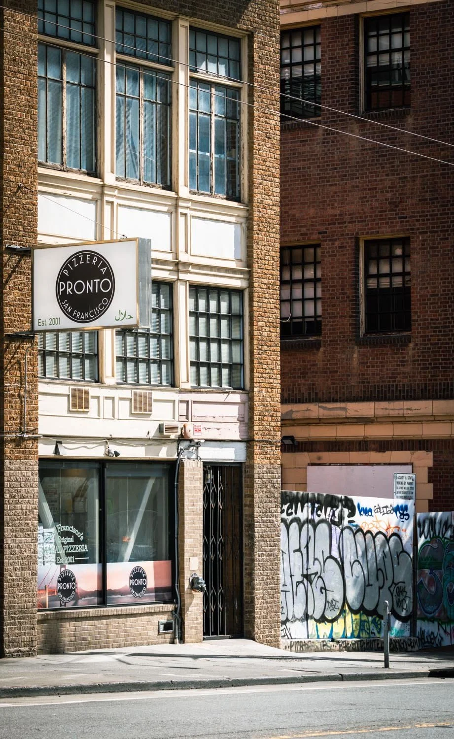 A street view of a brick building with large windows, a pizza restaurant sign that reads "Pronto, San Francisco," graffiti-covered wall, and a sidewalk in front.