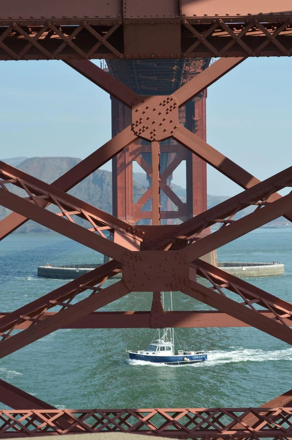 Underneath view of a red bridge with a sailboat sailing below, mountains in the background.