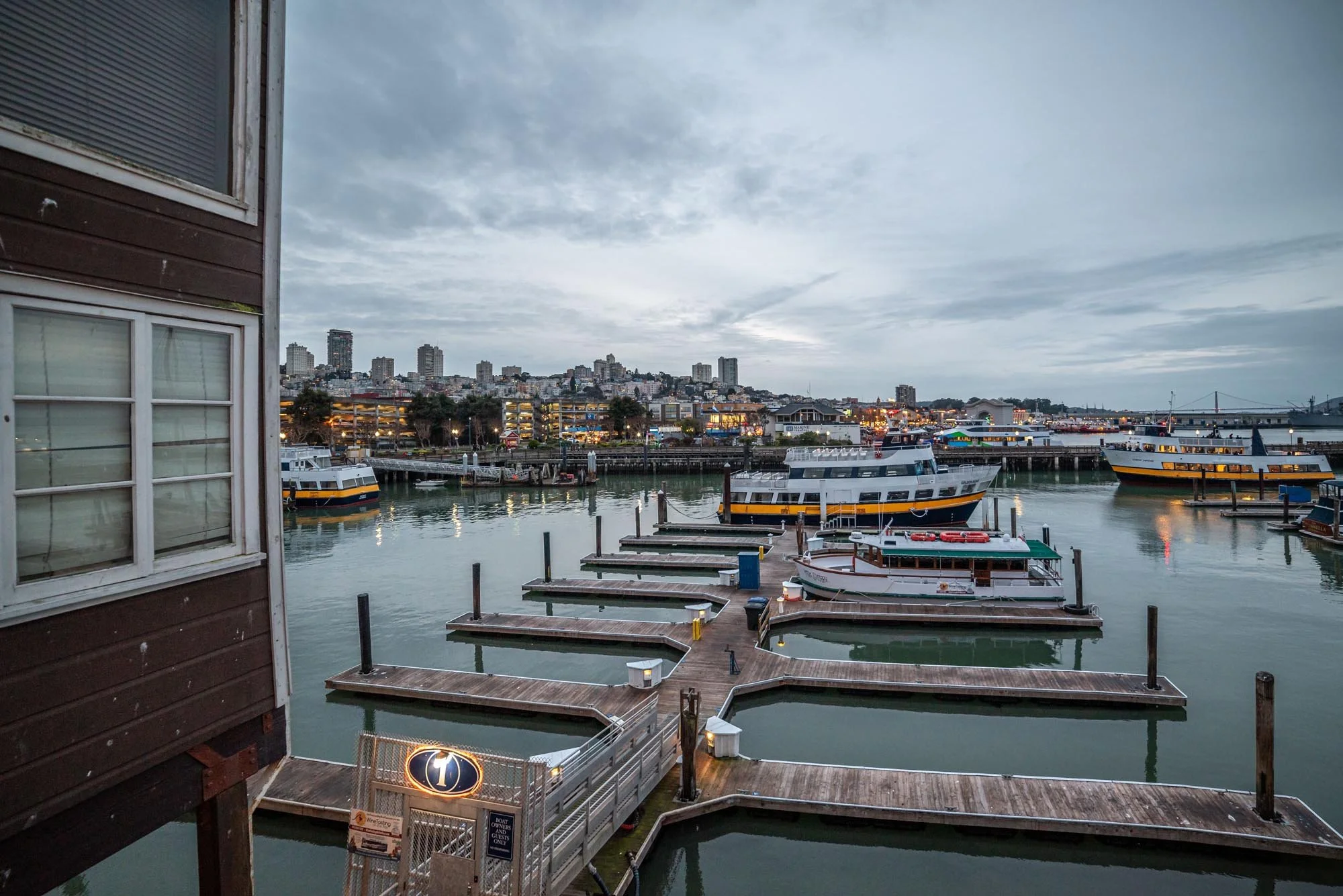 A view of a waterfront marina with boats docked at wooden piers, city buildings in the background, and a cloudy sky overhead.