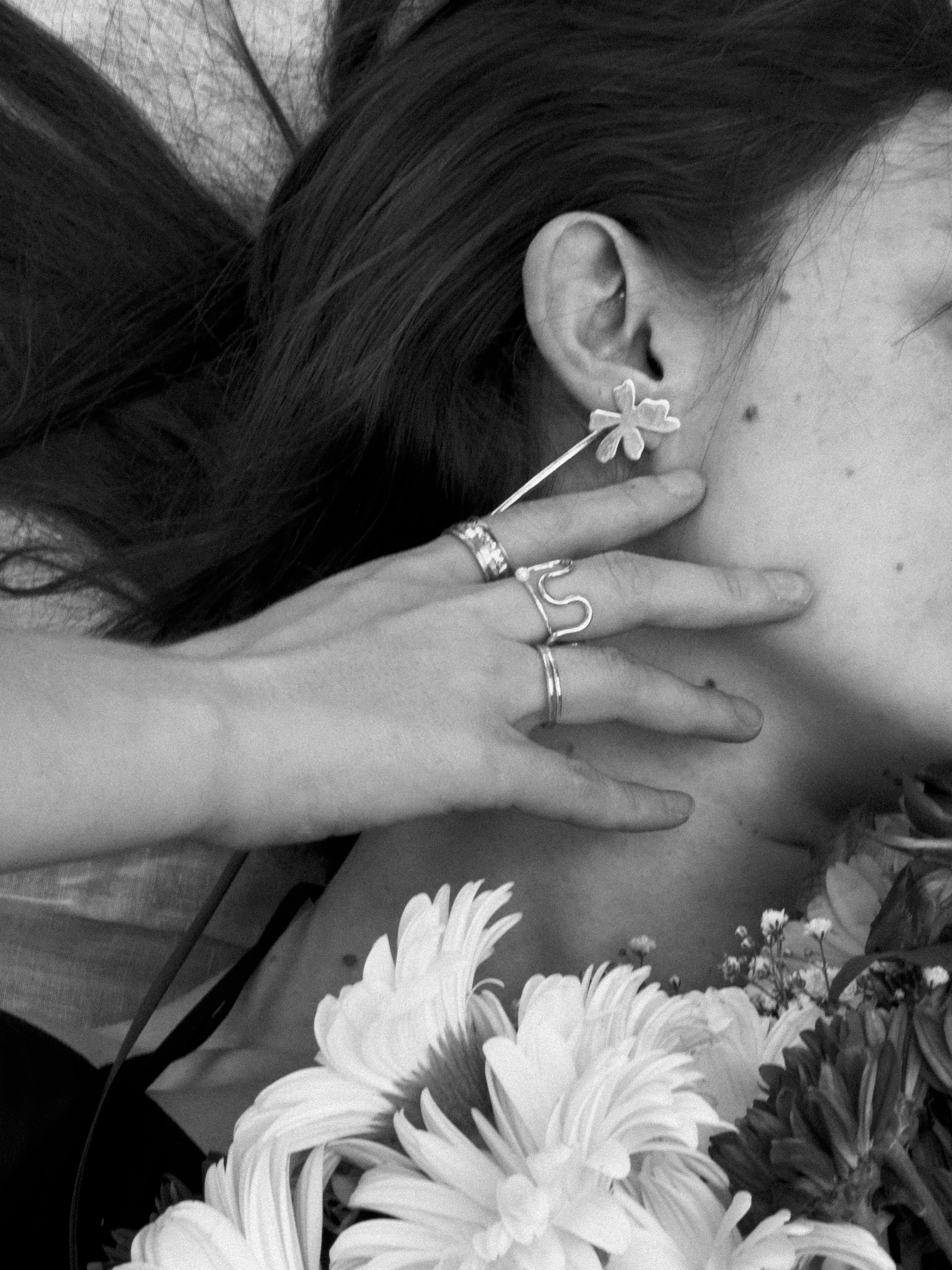 Primer plano de una mujer con pendientes de flor, tocándose la oreja y rodeada de flores.