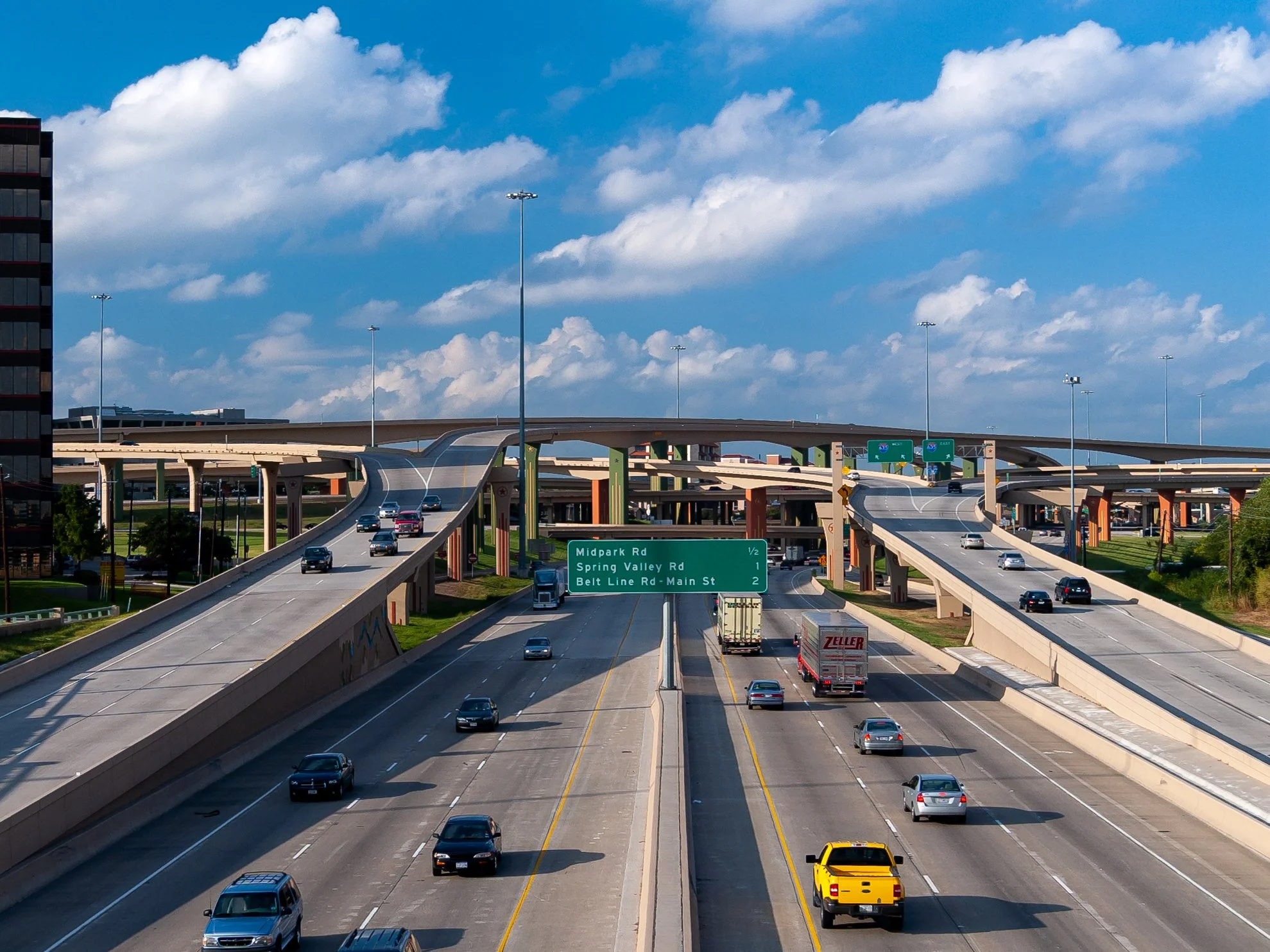 Multiple overpasses and highways with cars, trucks, and trucks driving on a clear day with blue sky and scattered clouds.