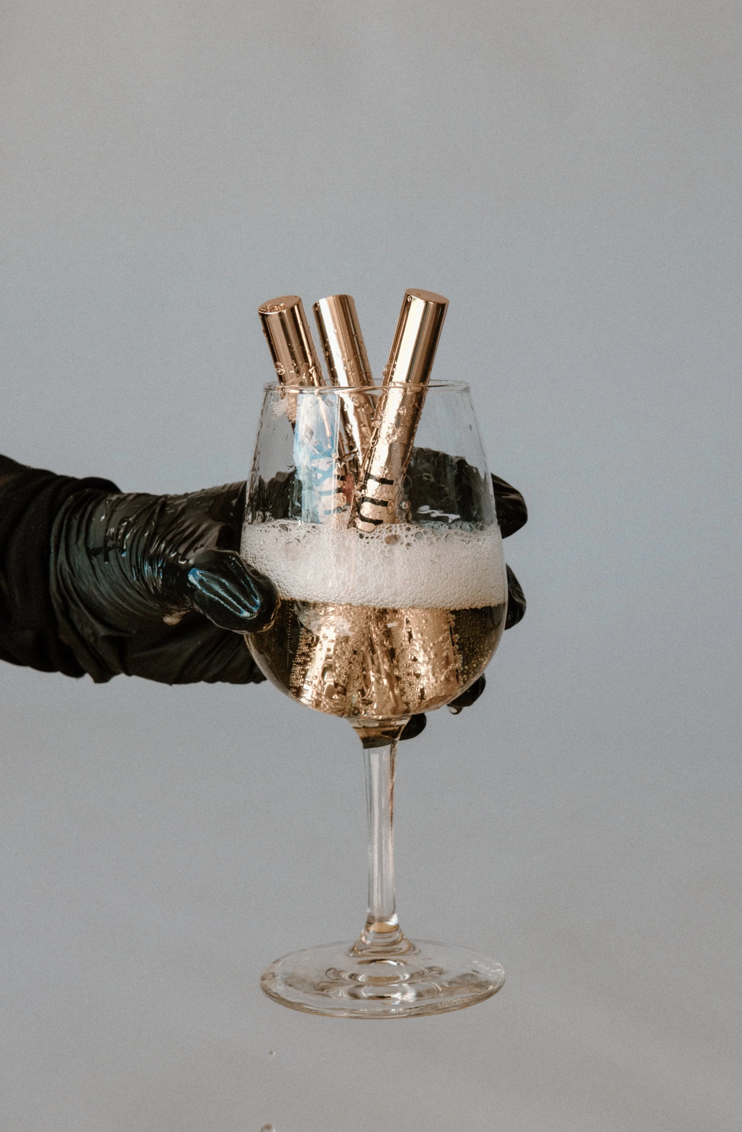 A hand wearing a black glove holding a wine glass filled with sparkling wine and three gold teeth whitening pens submerged in the bubbles.