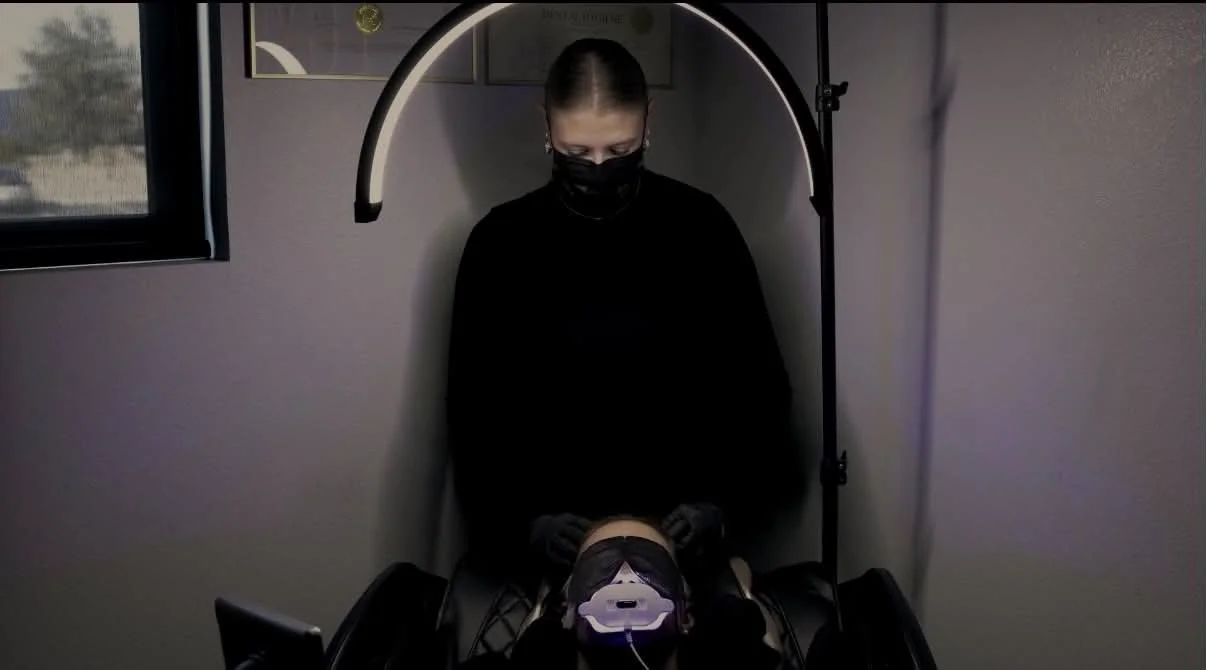 A woman wearing a mask and black clothes stands behind a patient lying in a full body massage chair, with a ring light positioned above them, in a clinical setting with a window on the left. Teeth whitening specialist is performing a scalp experience