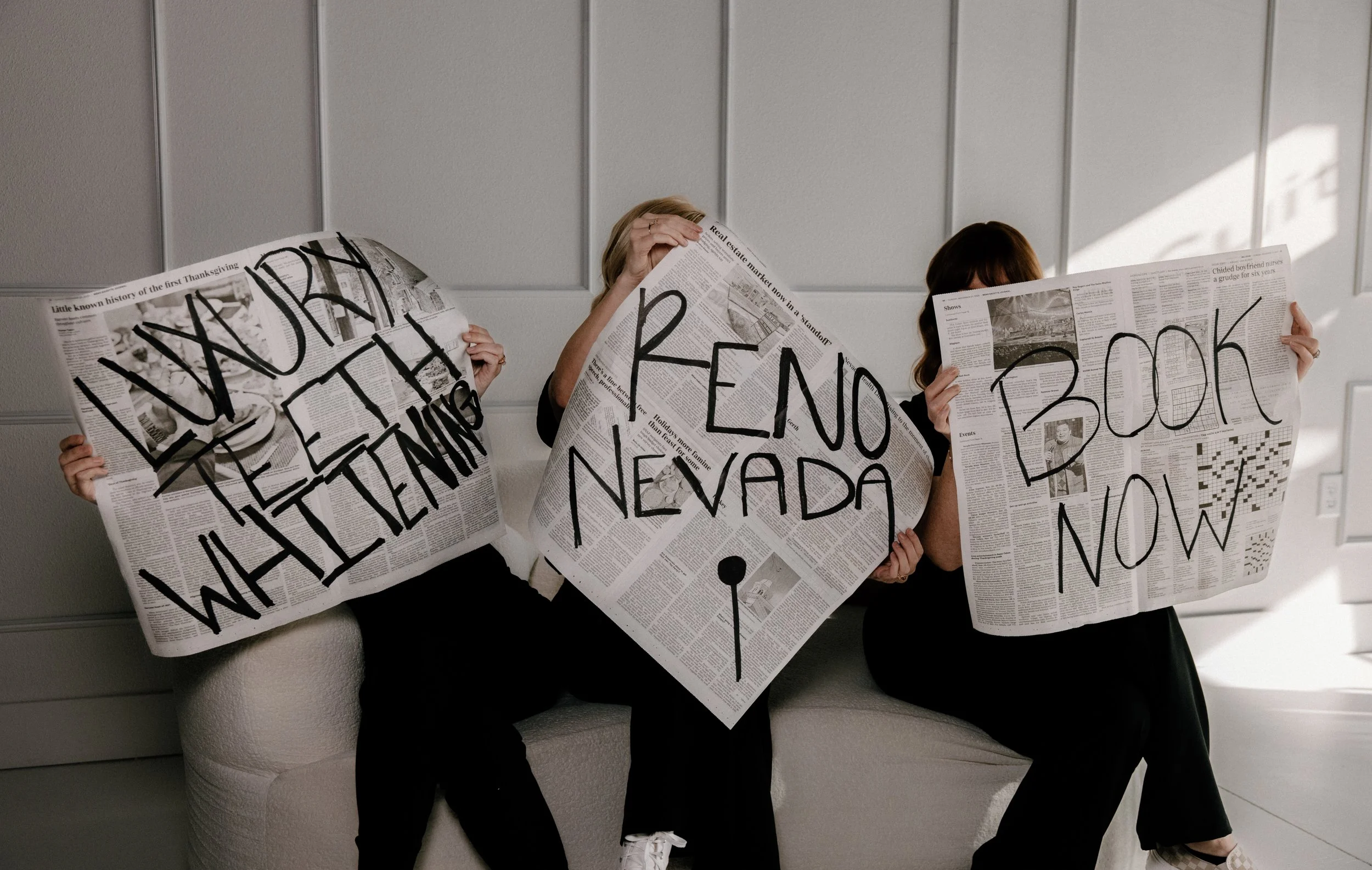 Three people sitting on a light-colored bench, each holding a newspaper with large handwritten words on the pages. The words say "NEVADA," "RENO," and "BOOK NOW." The background is a plain wall with vertical paneling, and sunlight is casting shadows on the wall.