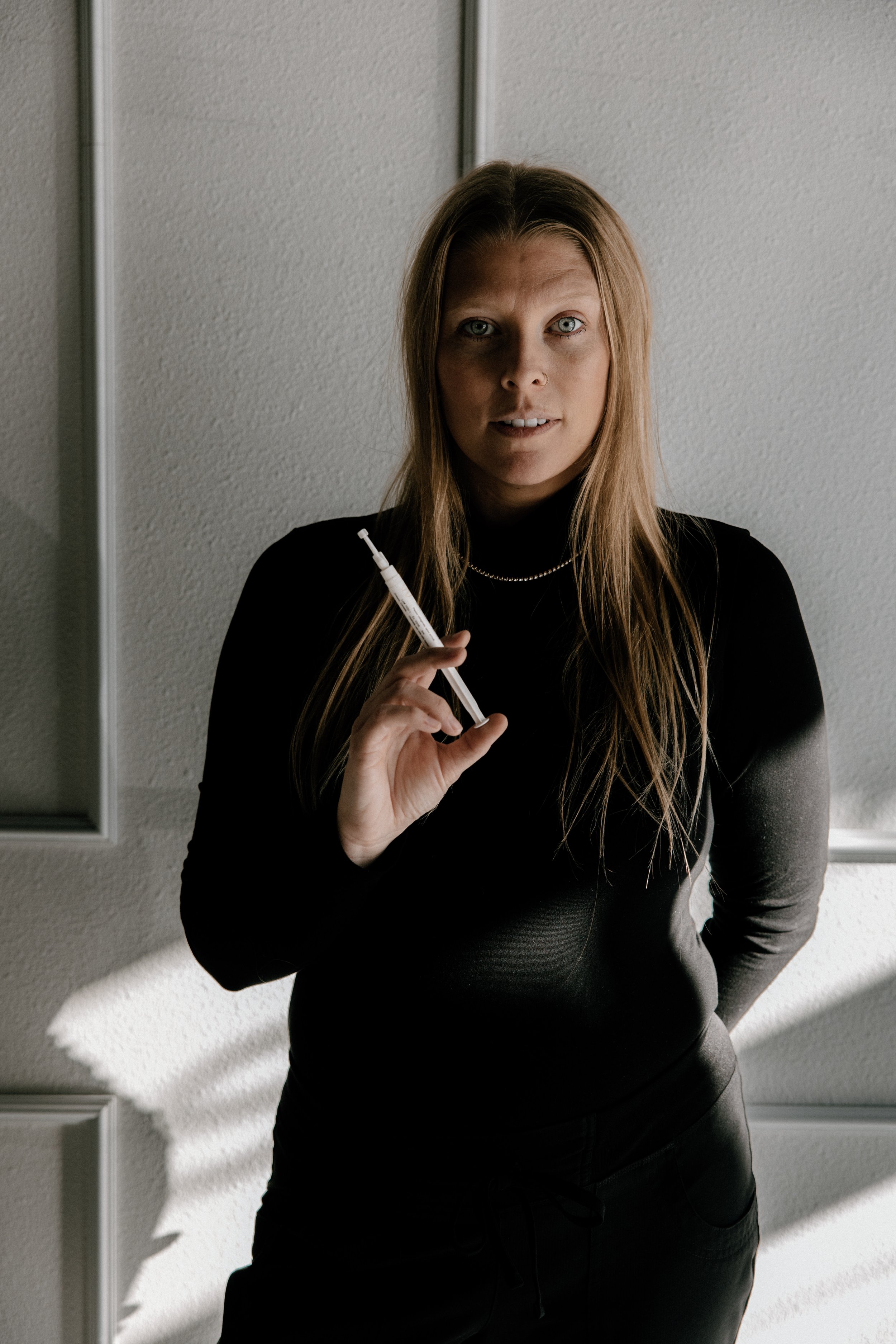 A woman with long blonde hair, light skin, and blue eyes, wearing a black top and pearl necklace, holds a teeth whitening syringe in her right hand and looks into the camera.