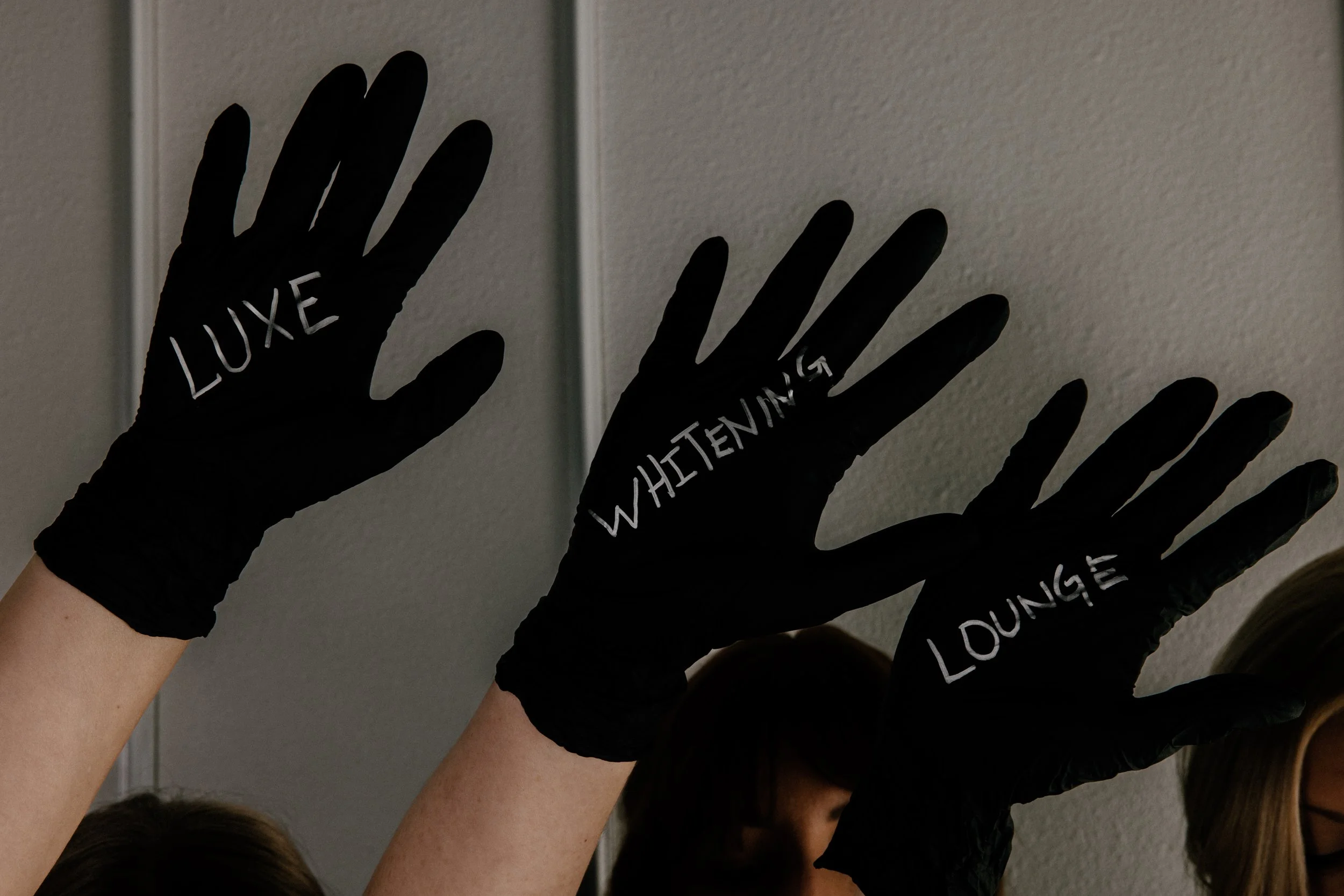 Three people wearing black gloves with silver handwritten words: 'LUXE', 'WHITENING', and 'LOUNGE', are raising their hands against a neutral background.