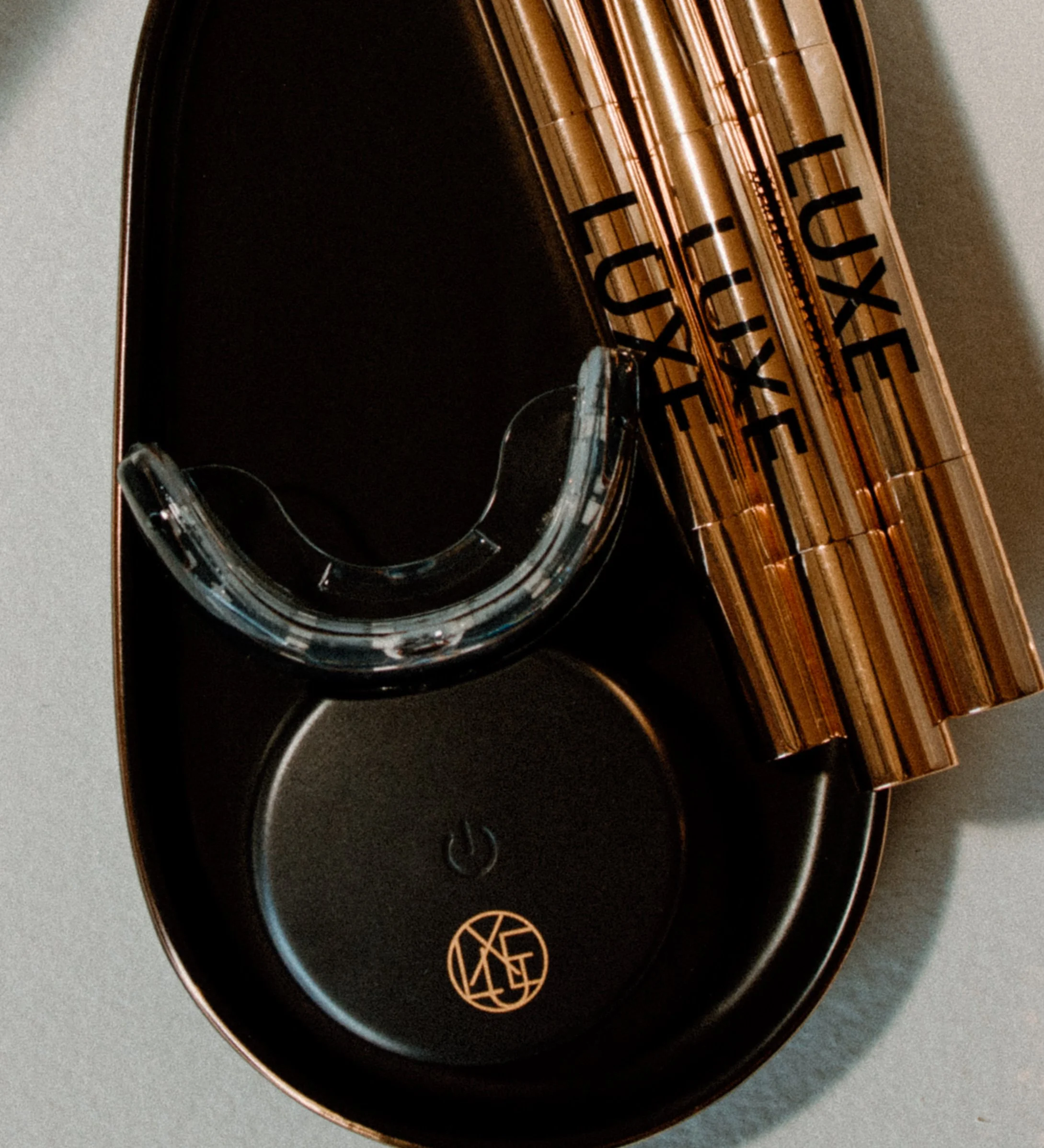 Close-up of a black tray with a black LED teeth whitening mouth piece with a gold symbol and text that reads 'LUXE' on a gray surface with 3 gold whitening pens.