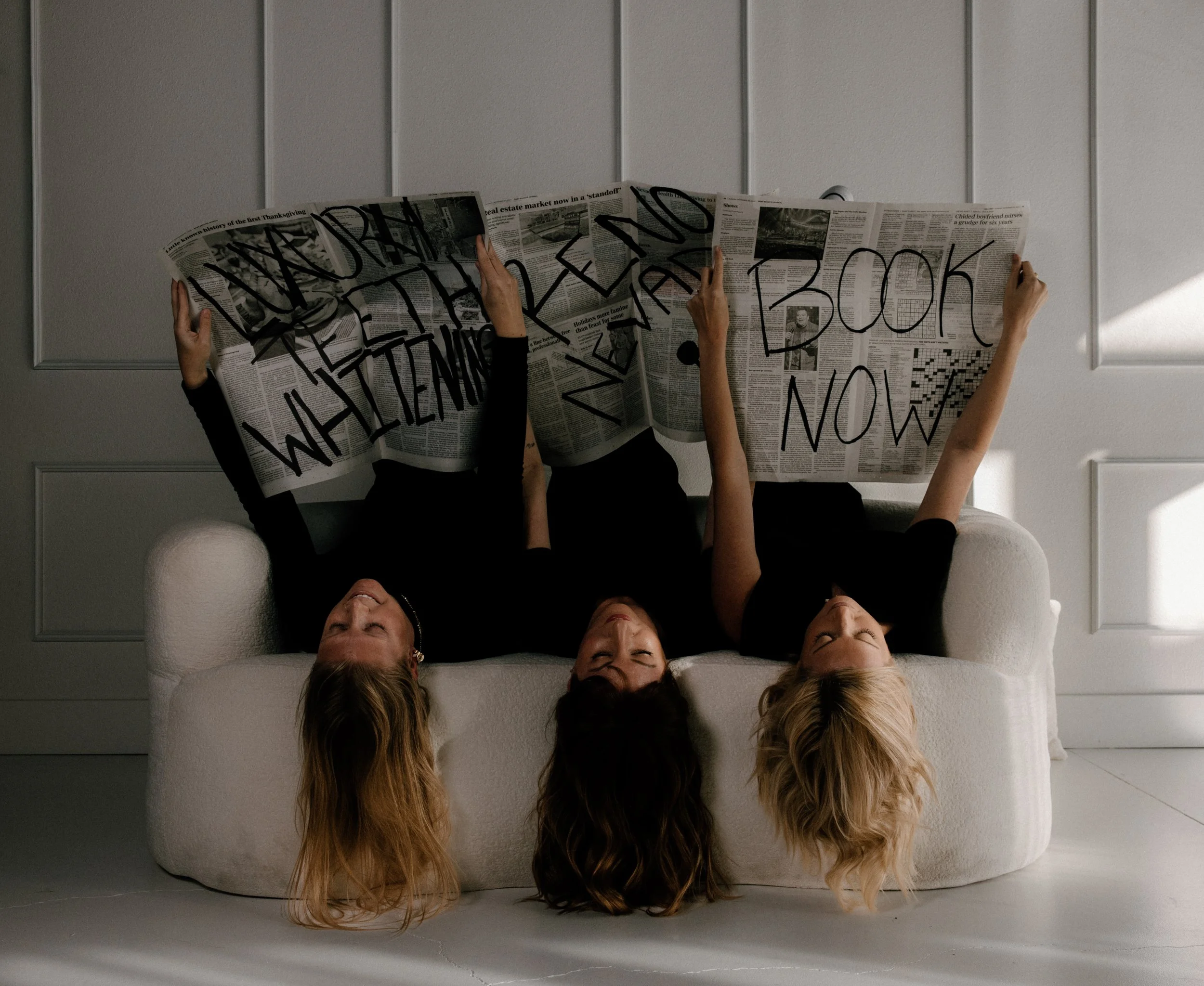 Three women with long hair lying upside down on a white sofa, reading newspapers with the words 'READING THE NEWSPAPER AND BOOK NOW' written in large black letters on the pages.