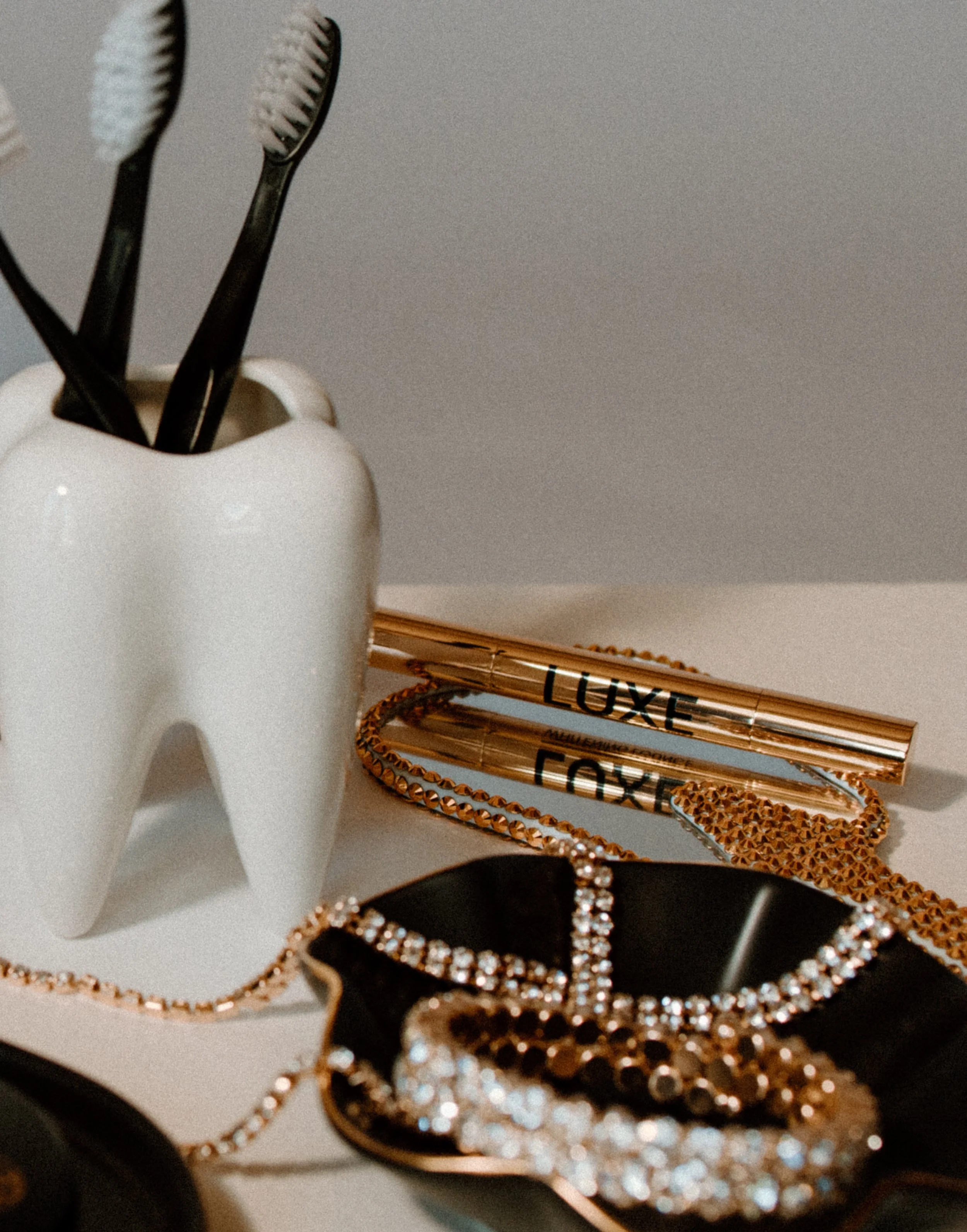 A white ceramic container holding three toothbrushes, a gold-colored whitening pens with the word 'LUXE' printed on it, a gold-colored bracelet, and a black jewelry holder.